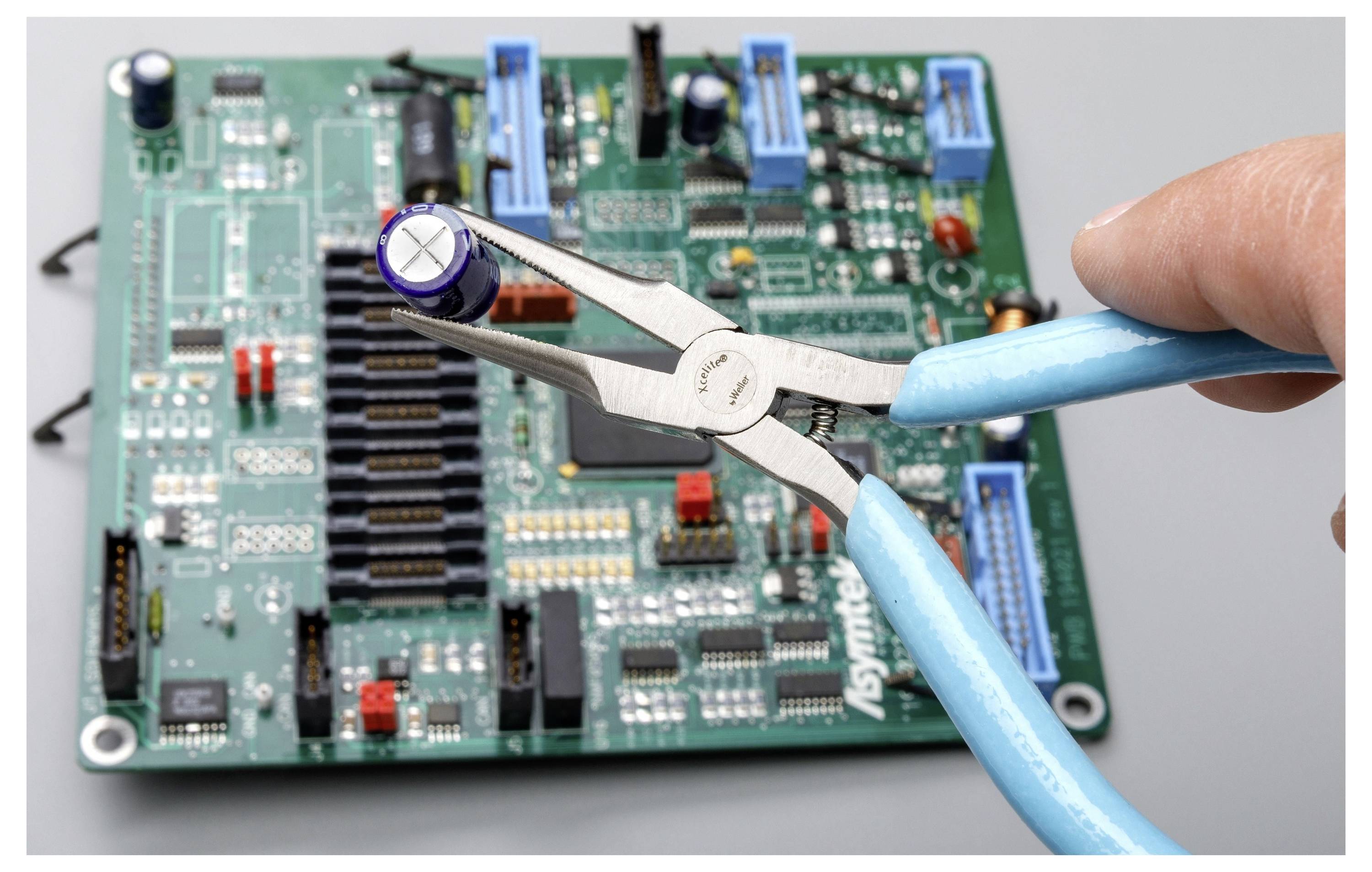 Nahaufnahme einer Zange, die einen Kondensator auf einer grünen Leiterplatte greift, was auf elektronische Reparatur oder Montage hindeutet.