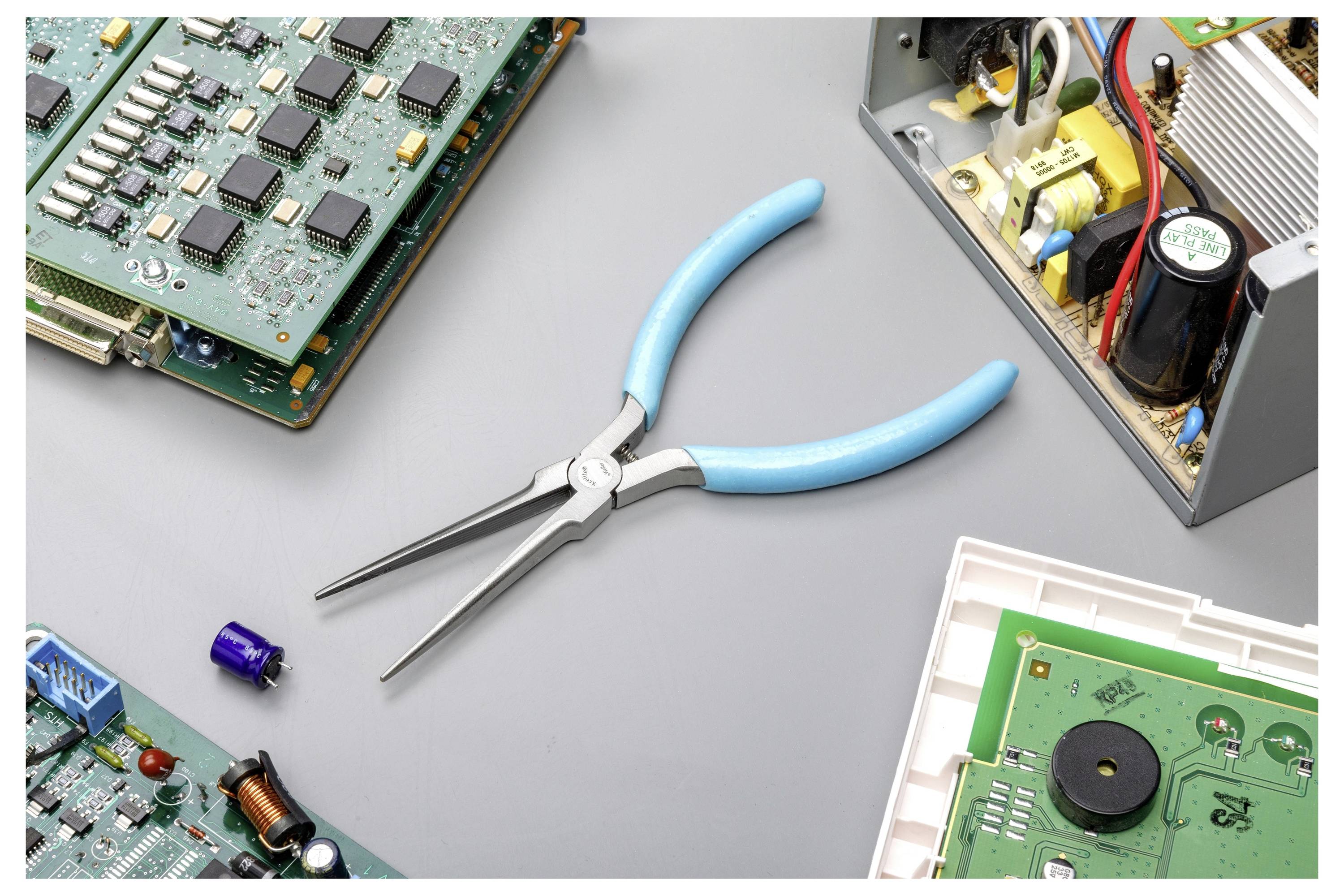 Blaugriffzange zentriert zwischen verschiedenen elektronischen Leiterplatten und Komponenten auf einer grauen Arbeitsfläche.