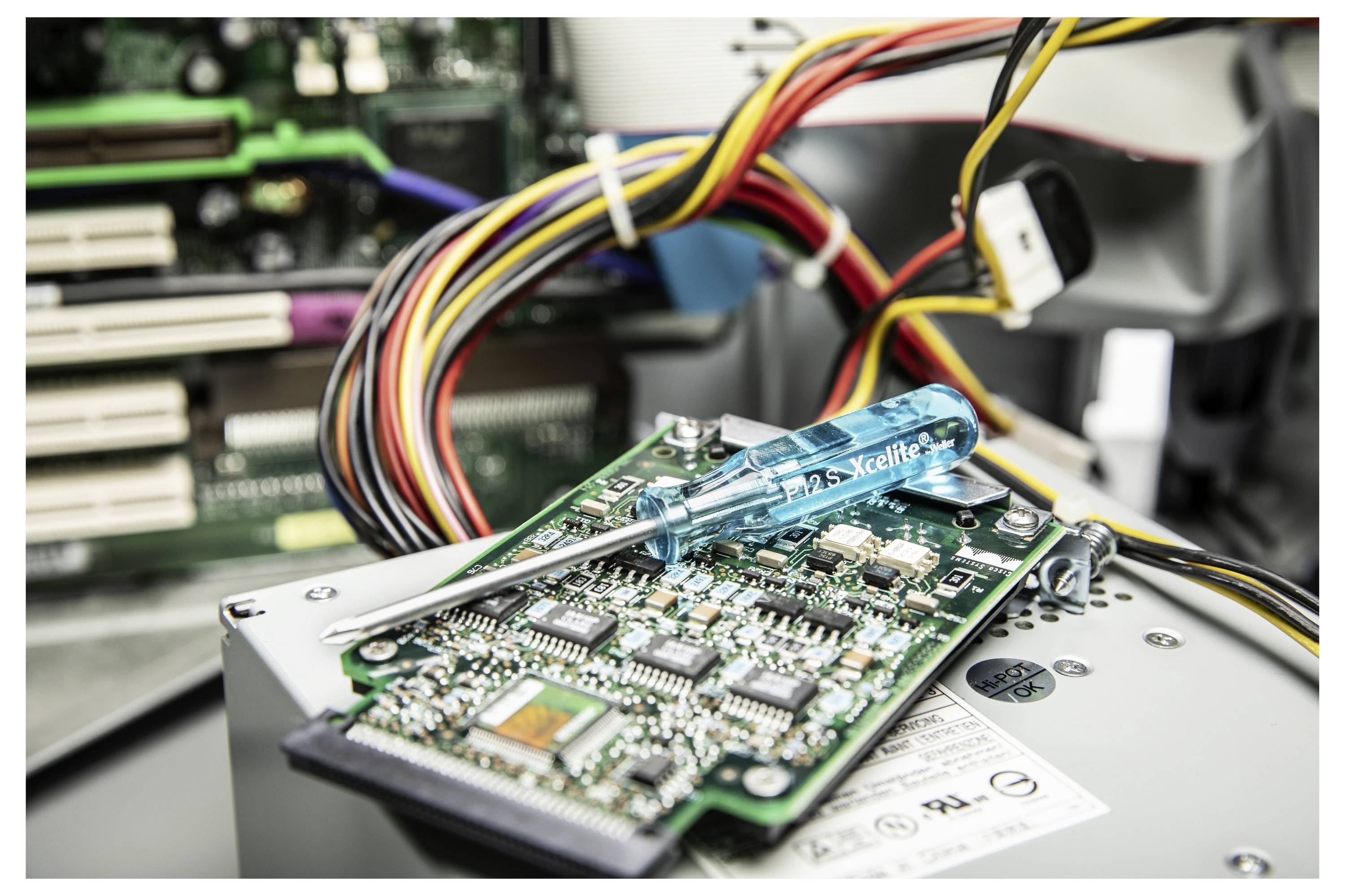 Nahaufnahme einer Computerfestplatte oder eines Schaltkreises, der repariert wird, mit einem blauen Schraubendreher, der auf dem Motherboard ruht, und gebündelten Kabeln im Hintergrund.