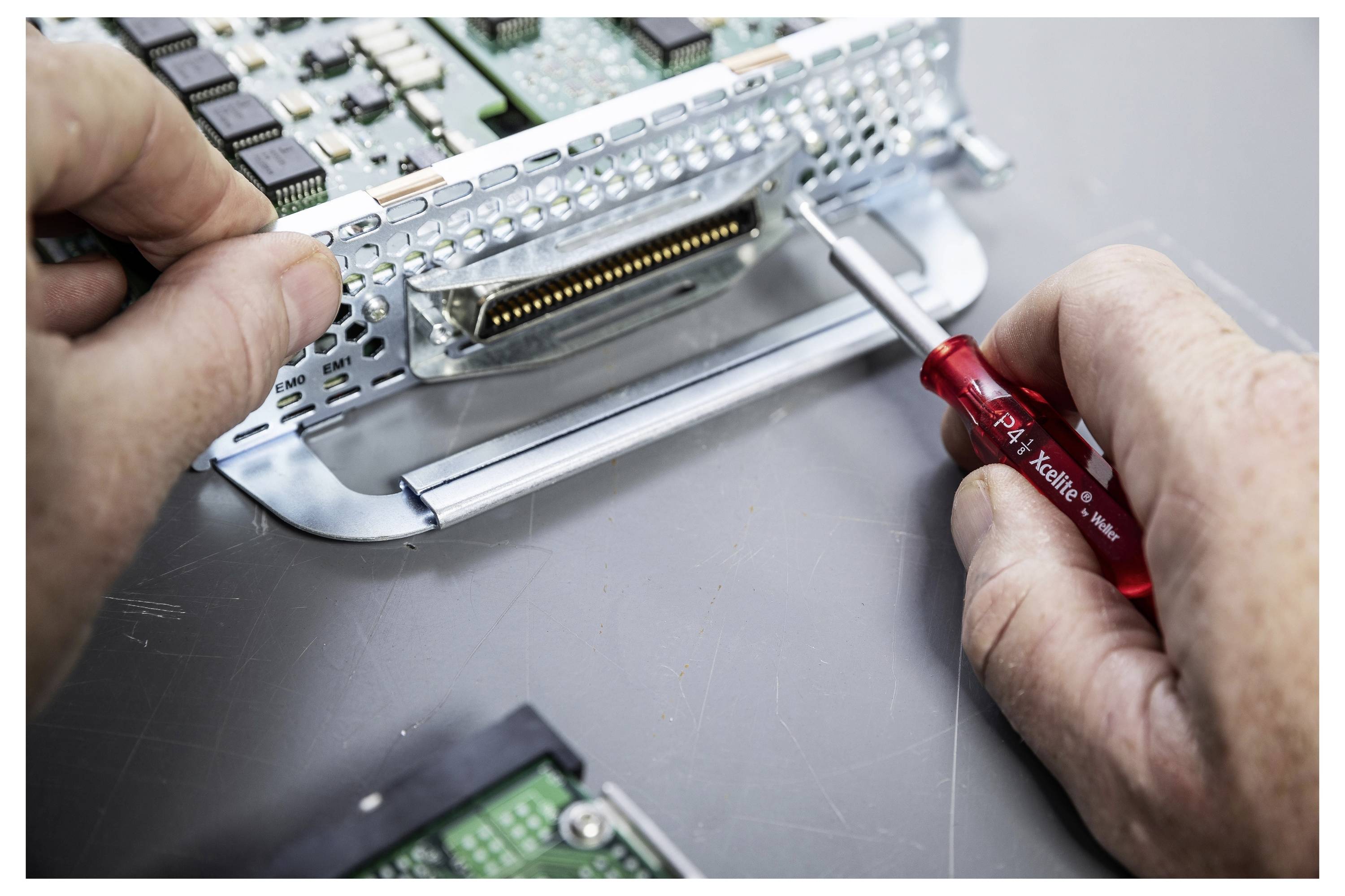 Hände verwenden einen kleinen Schraubenzieher, um ein metallisches elektronisches Bauteil oder einen Stecker in einem Gerät auf einer Werkbank einzustellen.