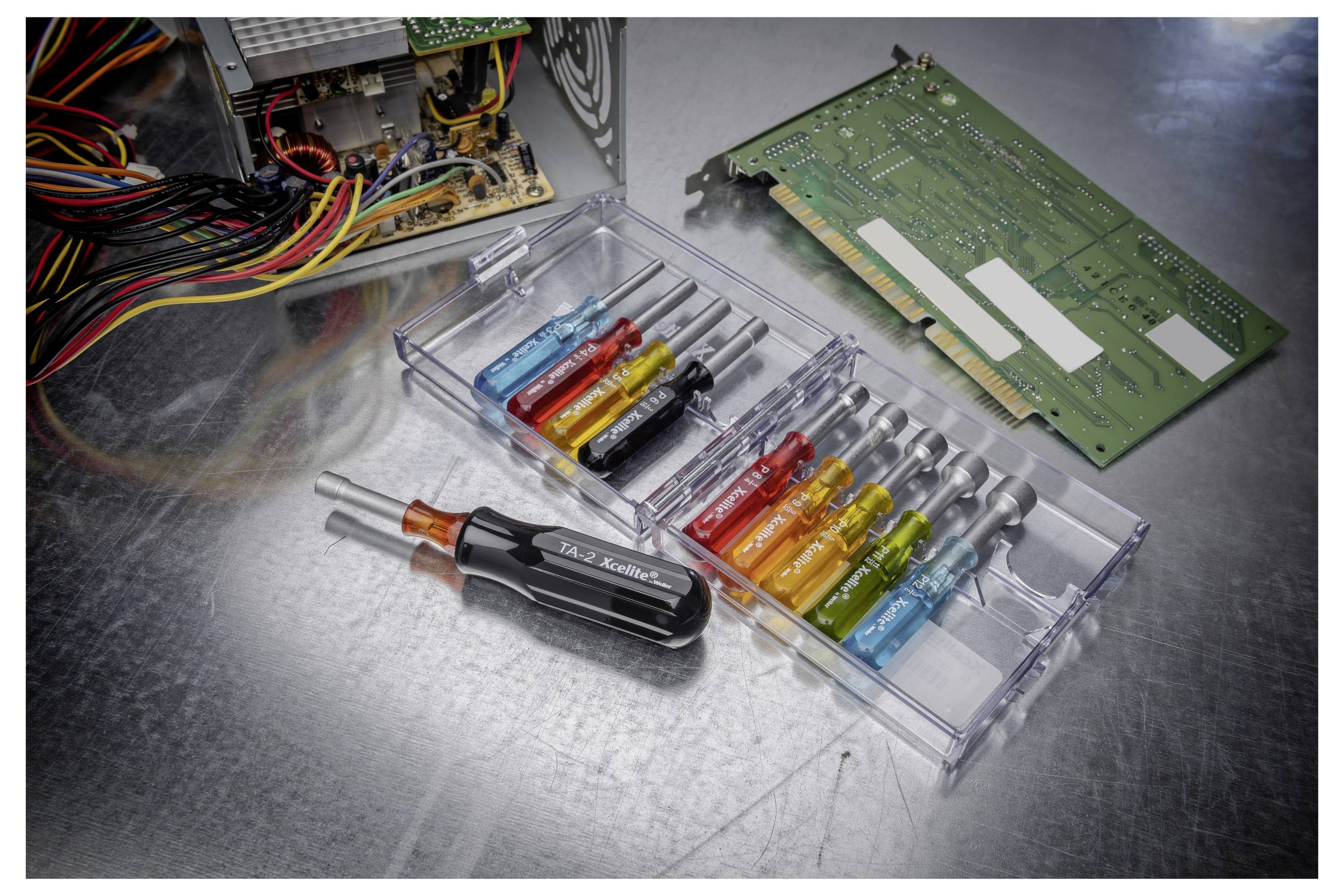 Ein offenes elektronisches Gerät mit freiliegenden Leitungen, einer Leiterplatte und einem durchsichtigen Behälter mit kleinen farbigen Schraubendrehern und Werkzeugen auf einer Metalloberfläche.