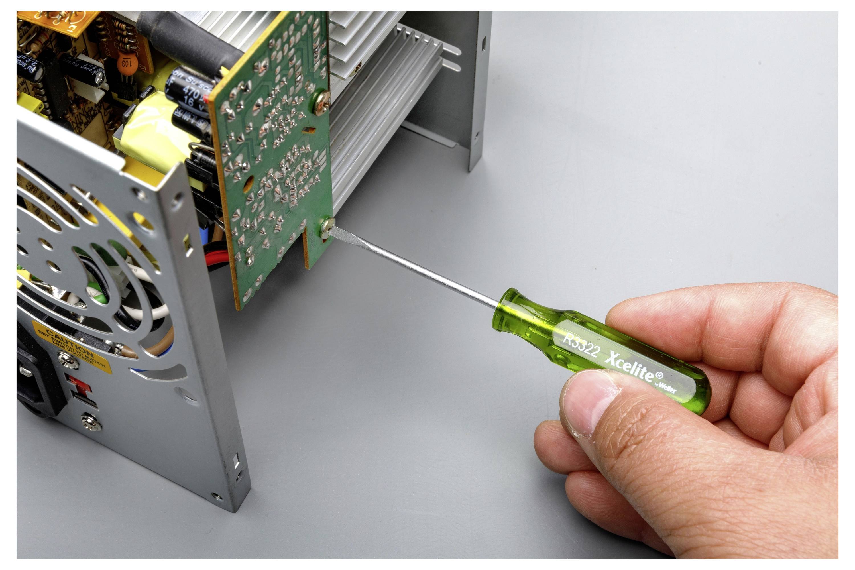 Eine Hand verwendet einen grünen Xcellite-Schraubendreher, um eine Schraube an der Seite eines elektronischen Geräts mit freiliegender Leiterplatte anzuziehen oder zu entfernen.