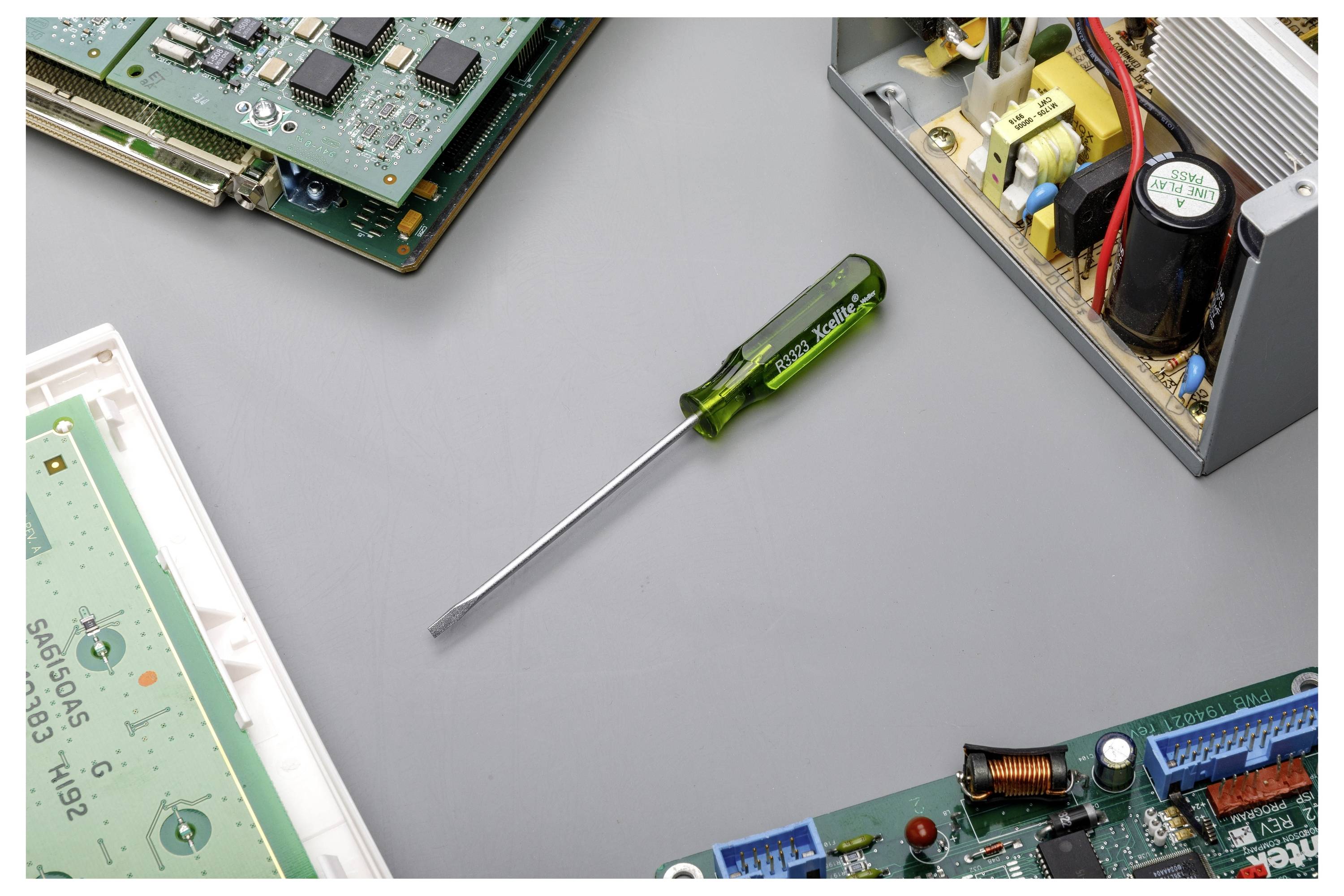 Grüner Schraubendreher mit Flachkopf auf einer grauen Arbeitsfläche, umgeben von zerlegten elektronischen Leiterplatten und einem Metallnetzteil.