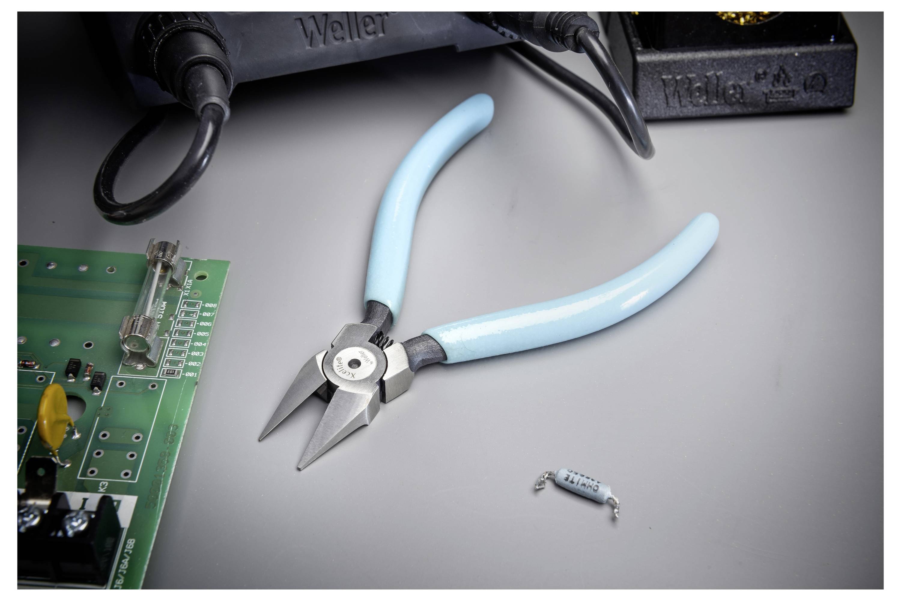 Elektronische Schneidezangen mit blauem Griff auf einer Werkbank neben einer Leiterplatte, einer Lötstation und einem kleinen Widerstand.