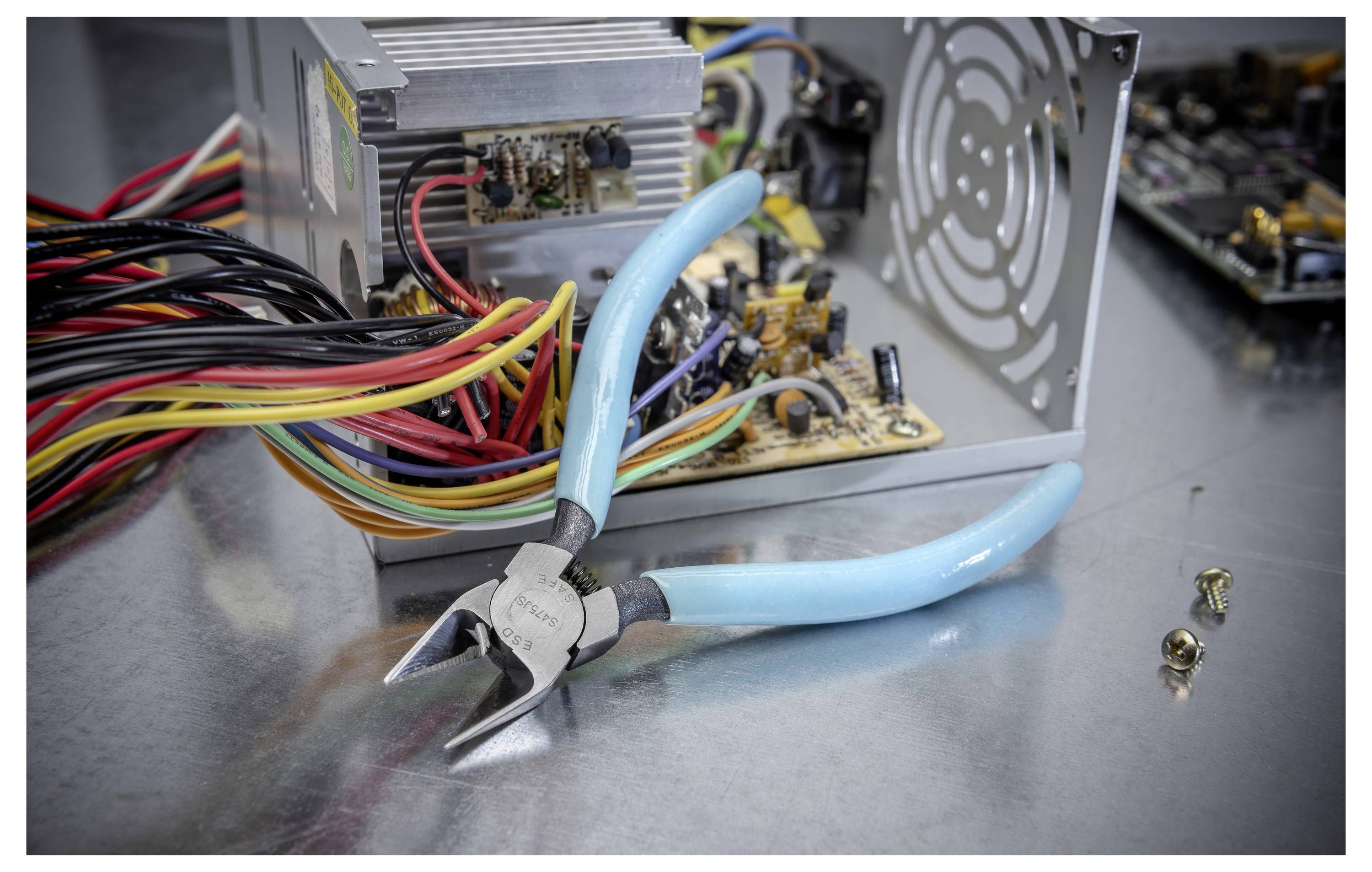 Blaugriffige Drahtschneider vor einem geöffneten Computernetzteil mit freiliegender Leiterplatte und gebündelten Kabeln auf einer Metalloberfläche.