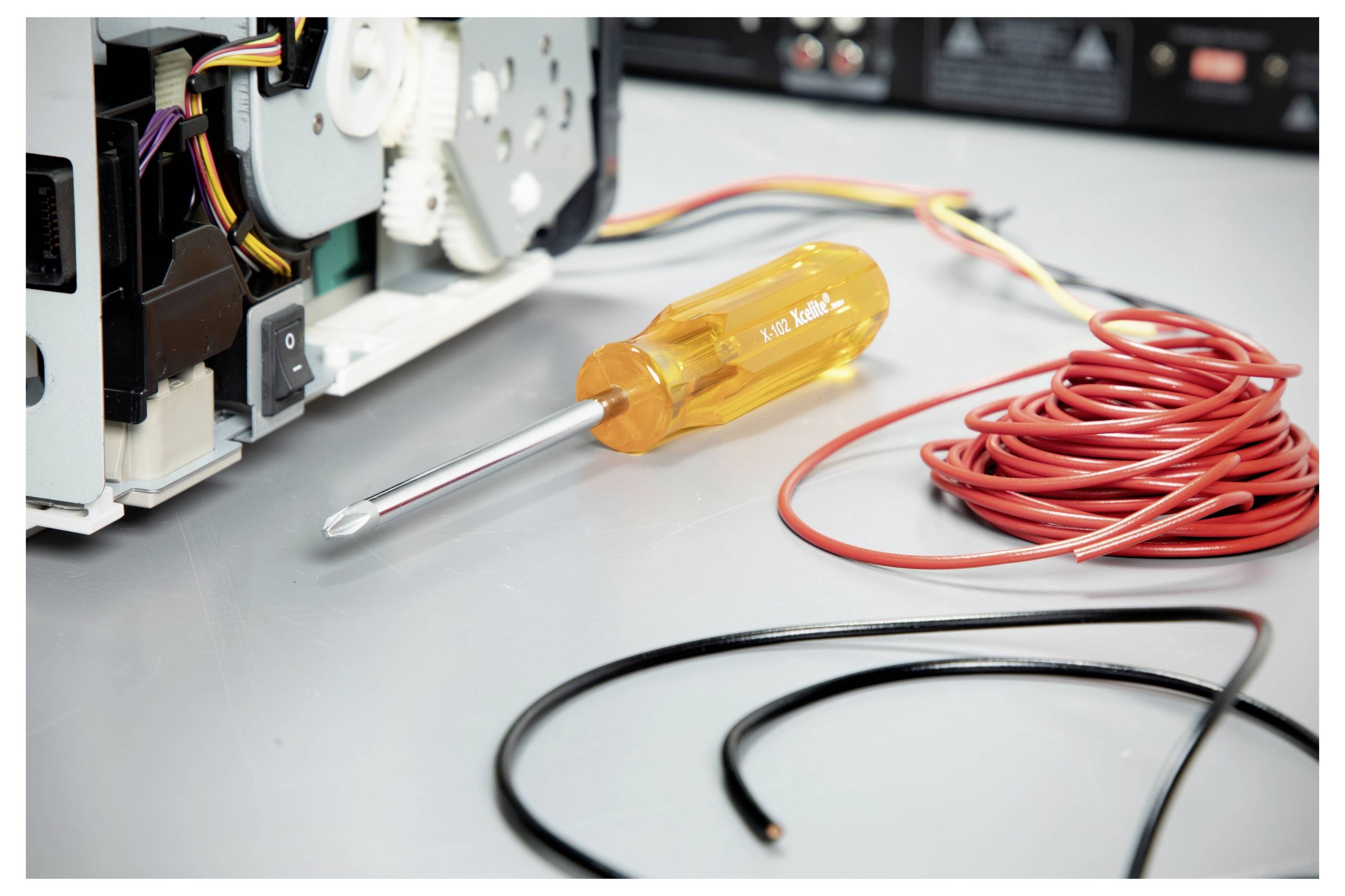 Schraubendreher neben einem offenen Computergerät mit aufgewickeltem rotem Kabel und lockeren Kabeln auf einer Arbeitsfläche, was auf Reparatur oder Montage hindeutet.