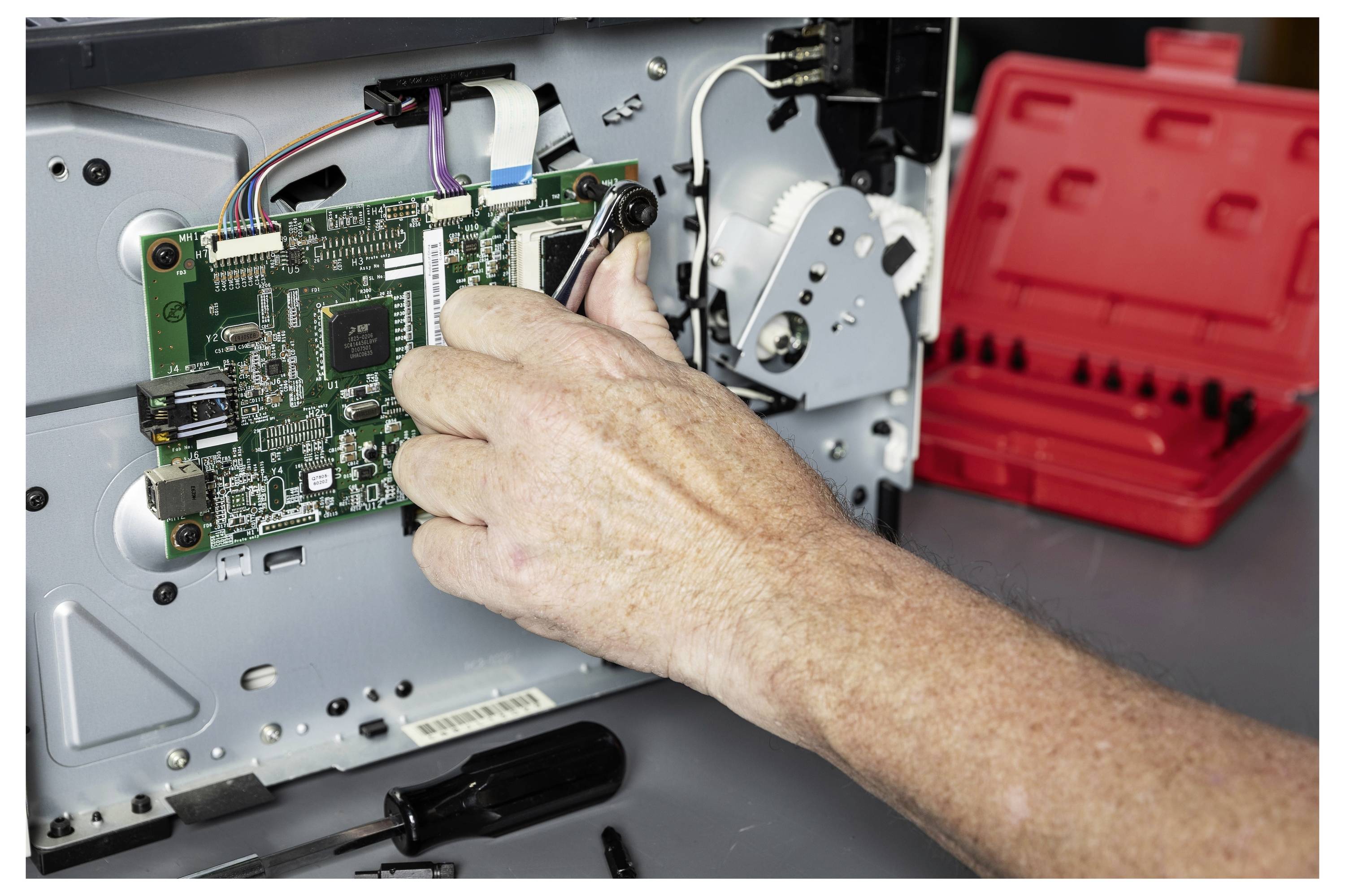 Eine Hand verbindet interne Kabel mit einer Leiterplatte in einem Computer oder Gerät während einer Reparatur, mit einem roten Werkzeugkasten im Hintergrund.