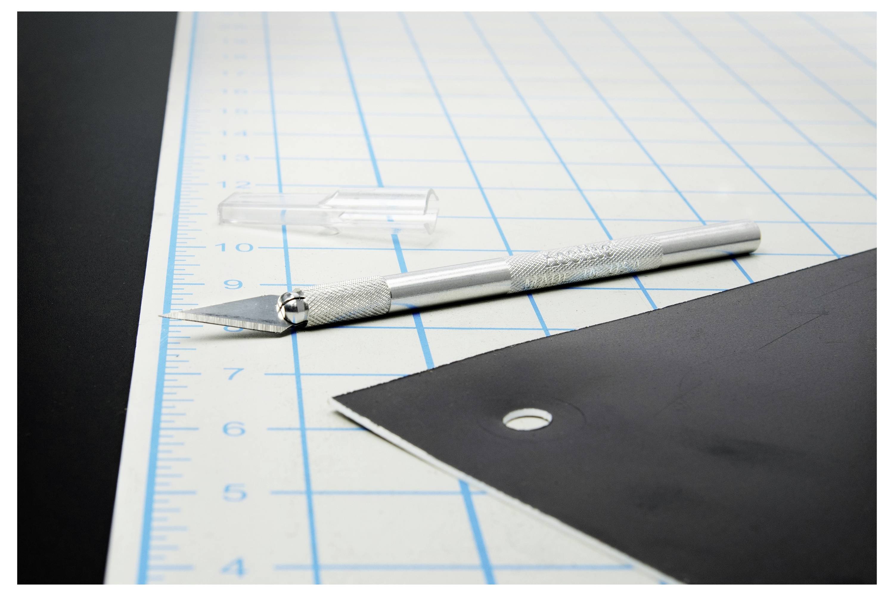Teppichmesser und Ersatzklinge auf einer Schneidematte neben einem schwarzen Blatt, was auf präzises Schneiden oder Handwerksarbeit hindeutet.