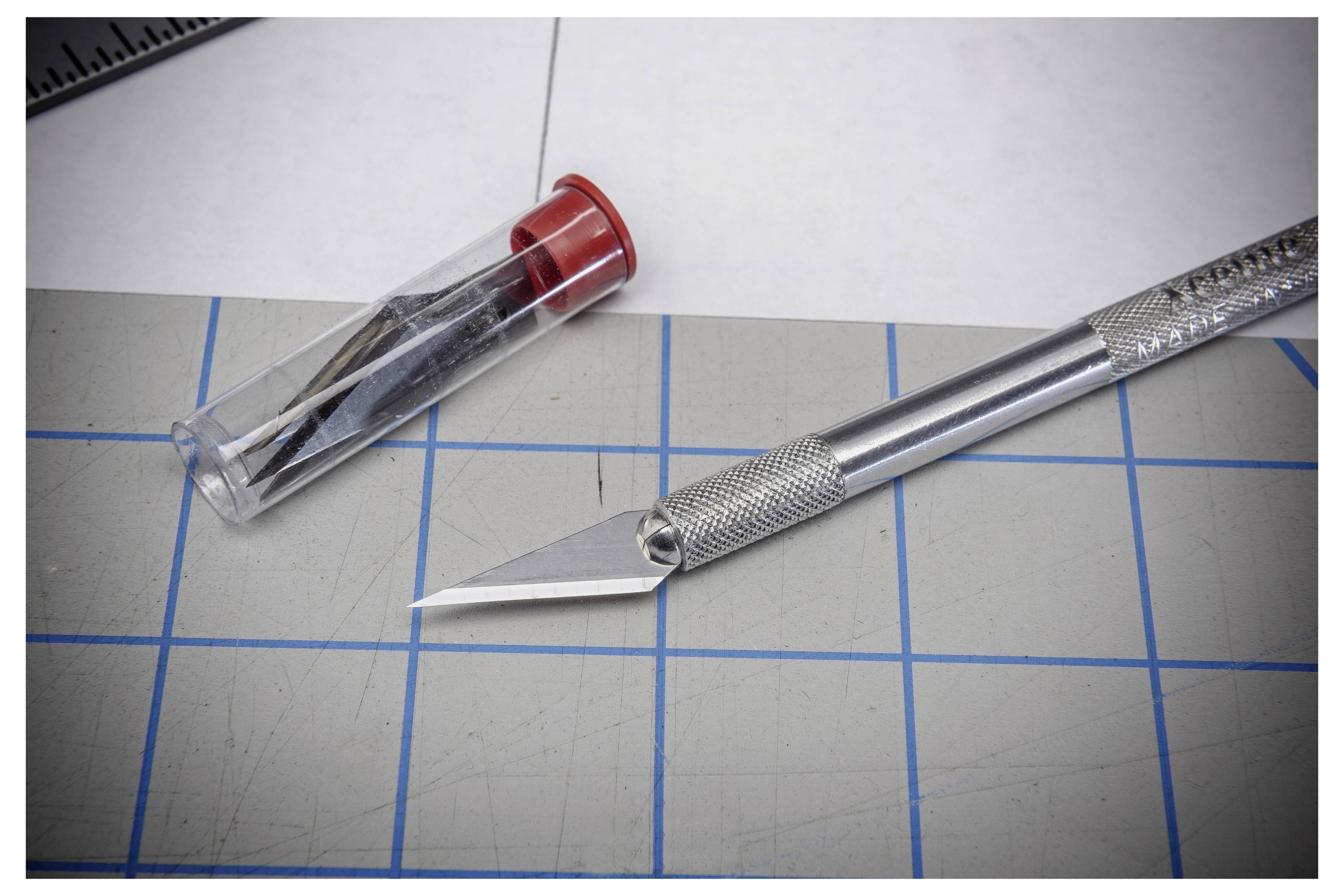 Ein X-Acto Bastelmesser mit Metallgriff und Klinge liegt auf einer karierten Schneidematte neben seiner durchsichtigen Kunststoffschutzkappe.