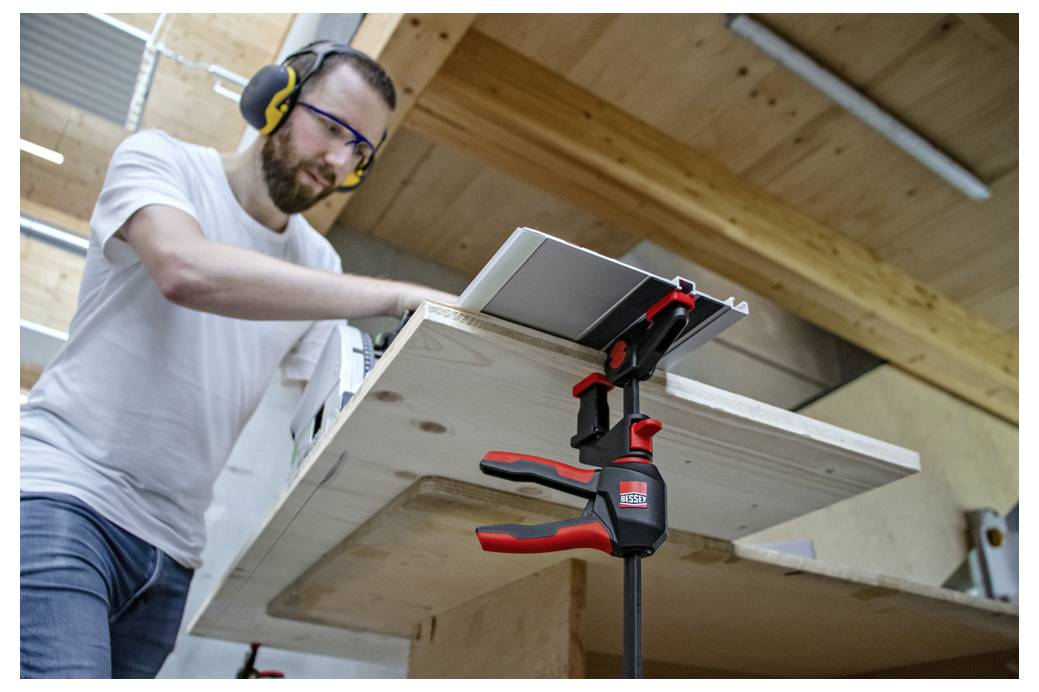 Ein Mann mit Schutzbrille und Gehörschutz spannt ein Holzbrett in einer Werkstatt auf einer Werkbank ein und verwendet ein Elektrowerkzeug zur Holzbearbeitung.