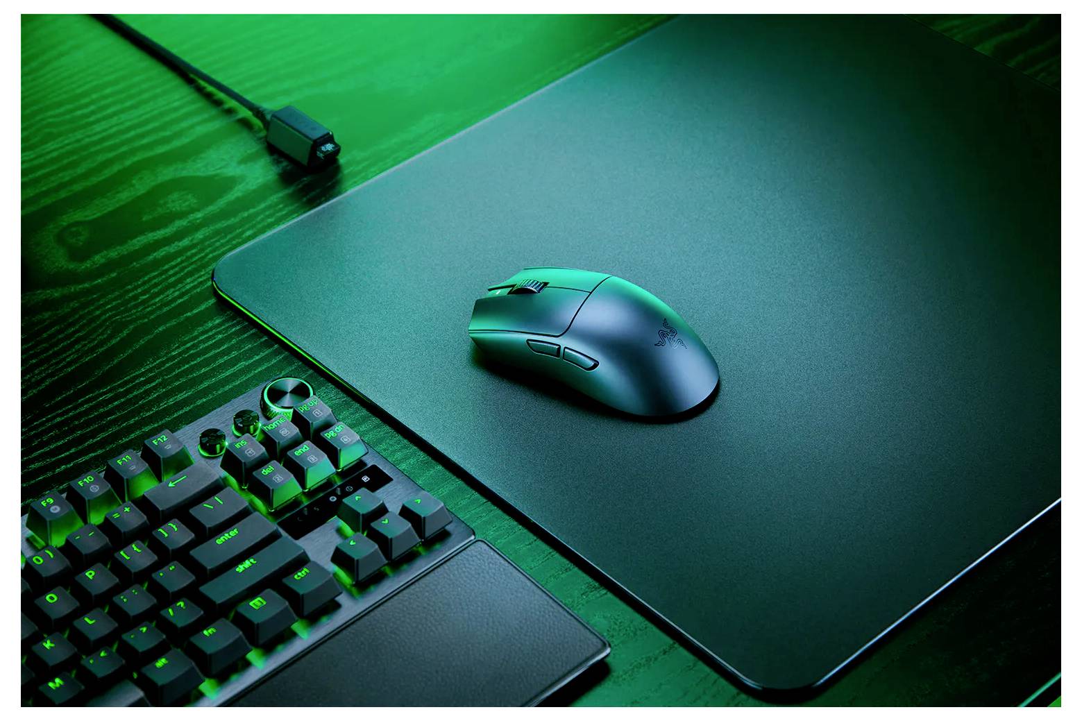 Eine kabellose Maus auf einem schwarzen Mousepad neben einer beleuchteten Tastatur auf einem Schreibtisch, die ein Computer-Setup zeigt.