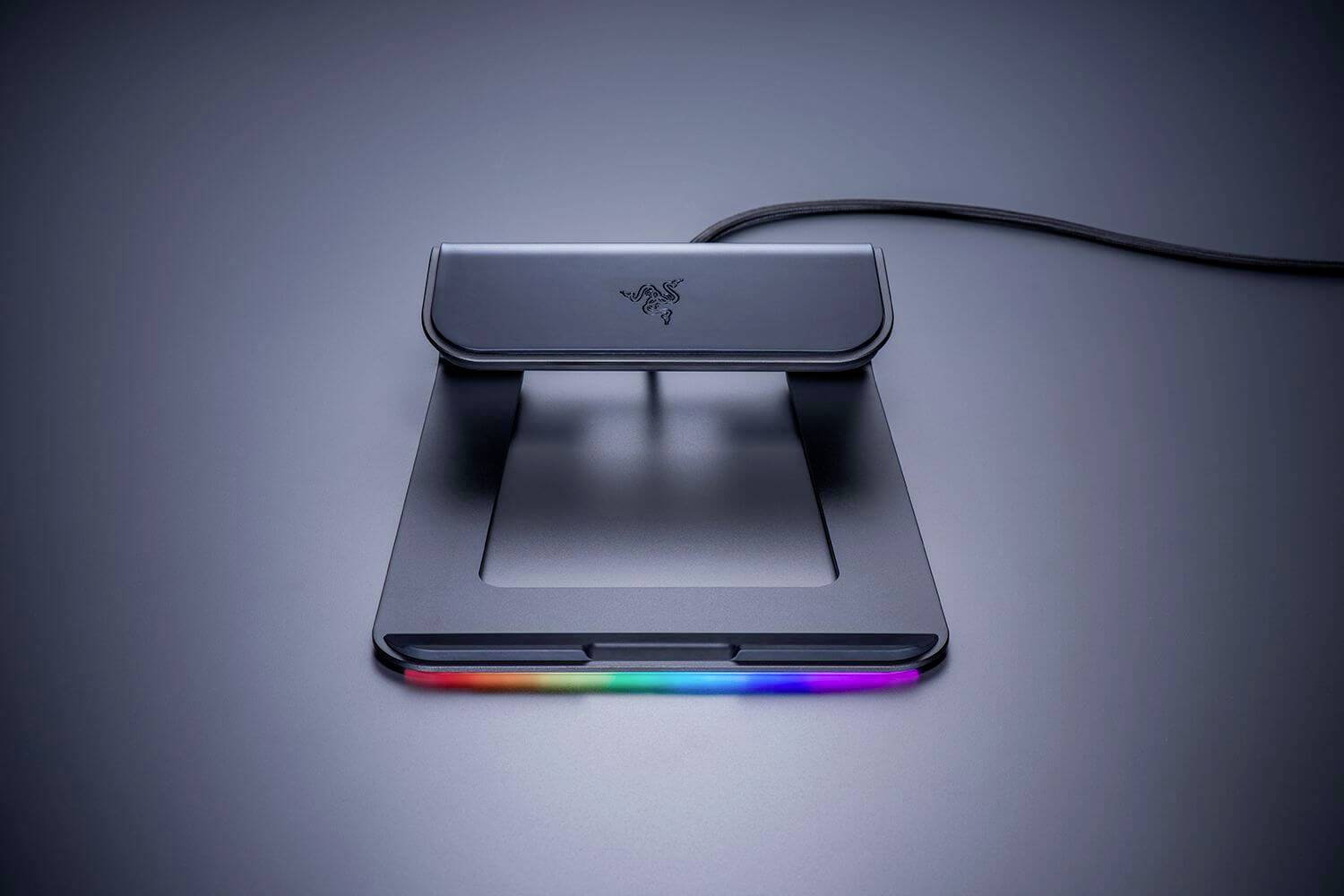 Ein schwarzer Laptop-Ständer mit einem Regenbogen-LED-Streifen entlang der vorderen Kante, gezeigt auf einer dunklen Oberfläche mit einem Kabel, das sich von der Rückseite erstreckt.