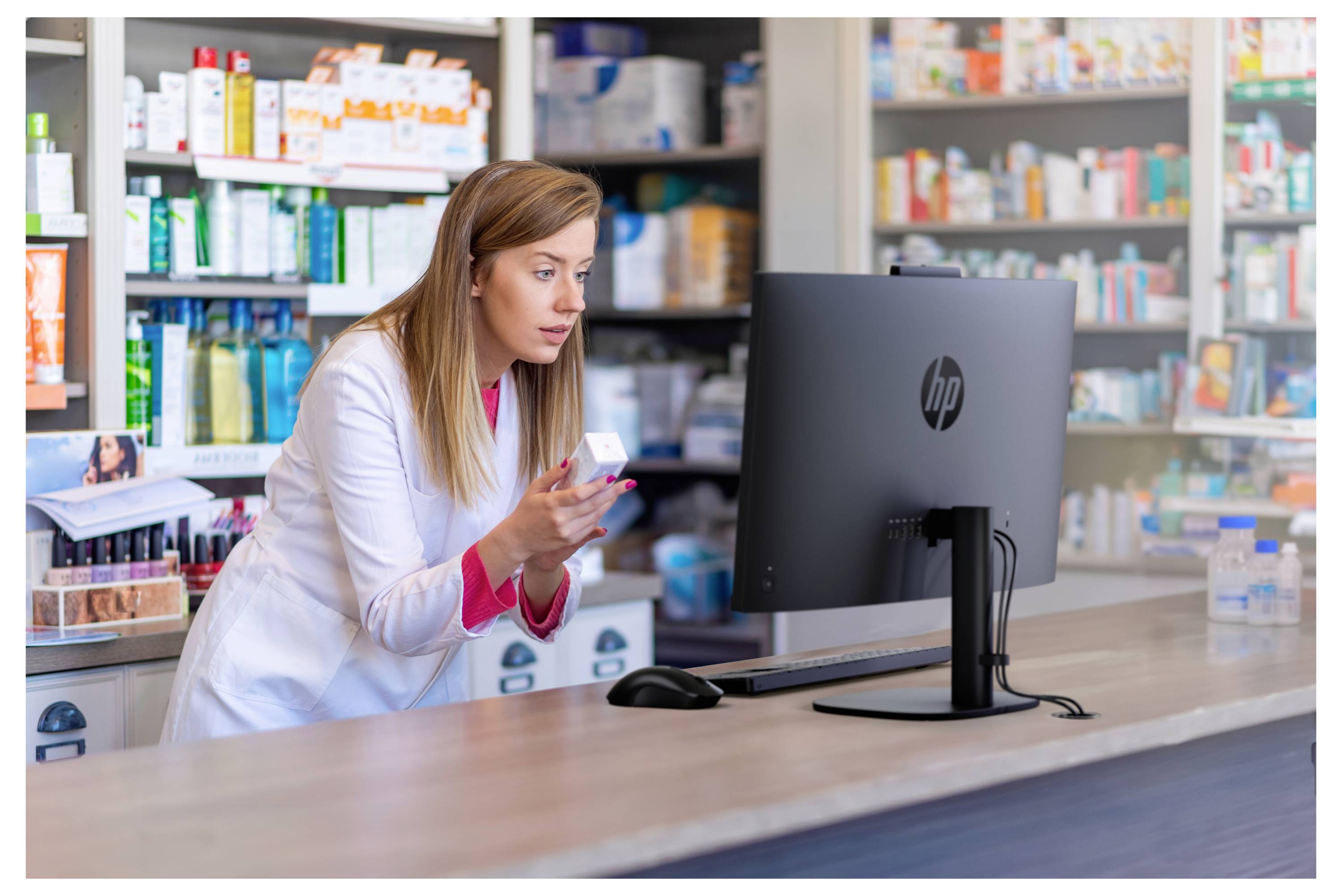 Apotheker in einem weißen Kittel prüft eine Medikamentenbox an einem Apotheken-Computer-Schalter, mit Regalen von Arzneimitteln im Hintergrund.