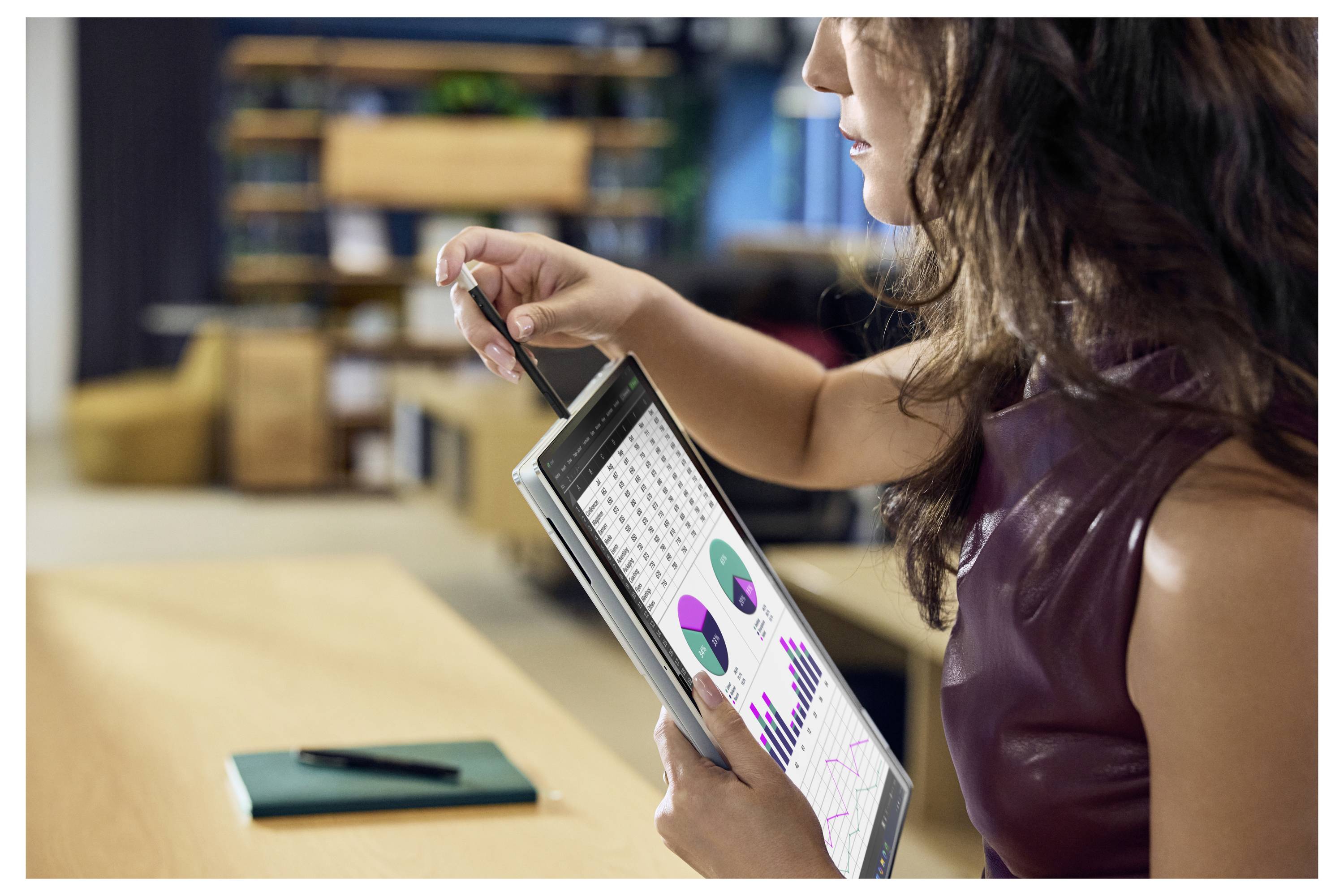 Frau in einem Büro hält ein Tablet mit Diagrammen und Grafiken, während sie darauf schreibt und neben einem Schreibtisch Daten überprüft.