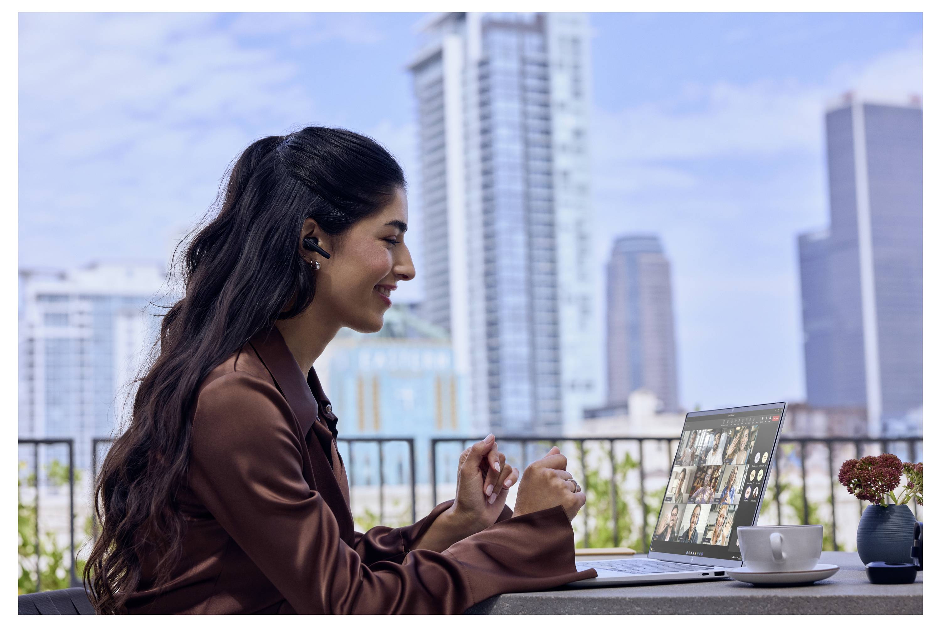 Frau in einer braunen Bluse führt ein Videogespräch auf einem Laptop in einem Outdoor-Café mit einer Stadtsilhouette im Hintergrund.
