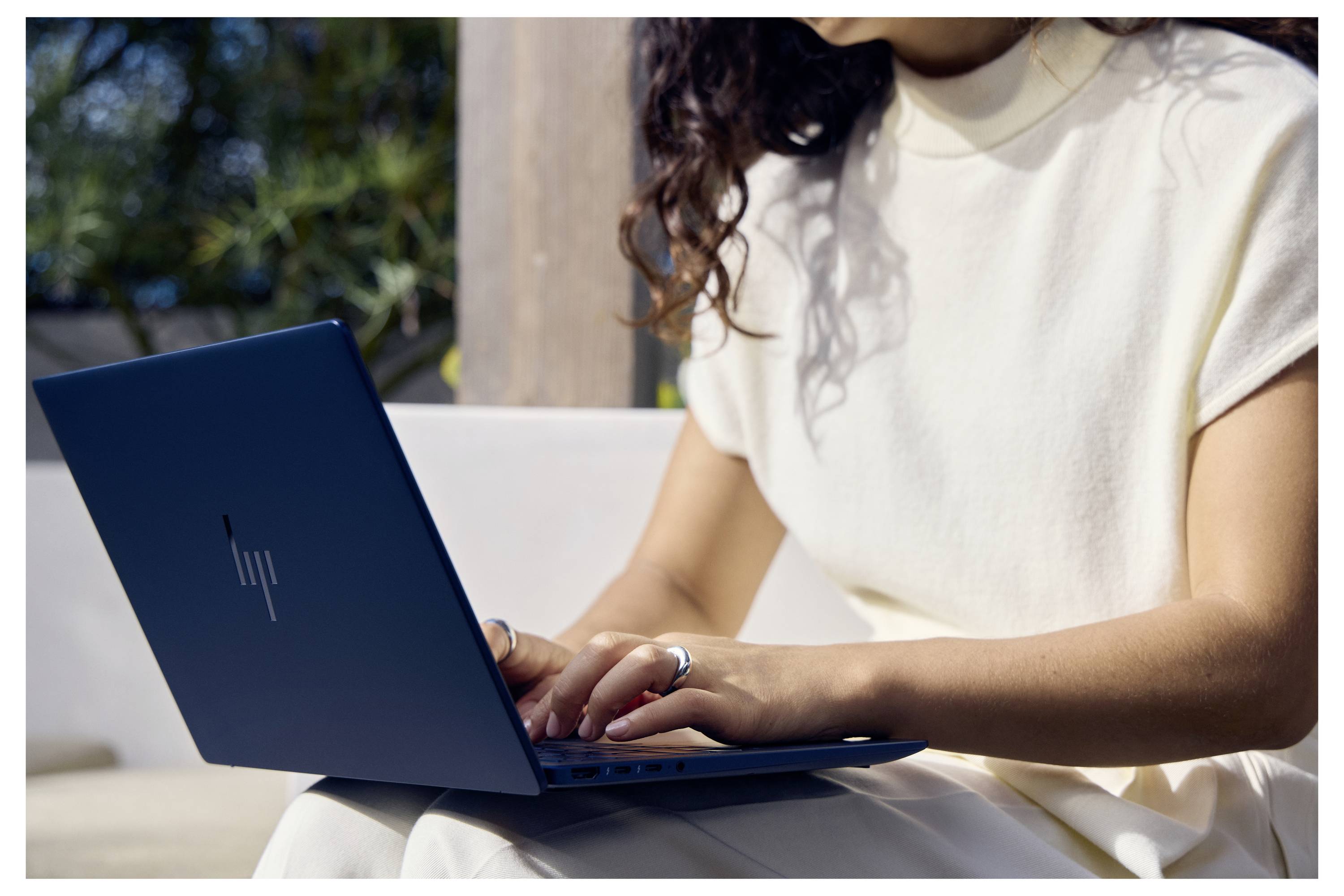 Eine Frau tippt auf einem Laptop im Freien bei Sonnenlicht, sitzt auf einem weißen Stuhl in einem lässigen cremefarbenen Outfit.