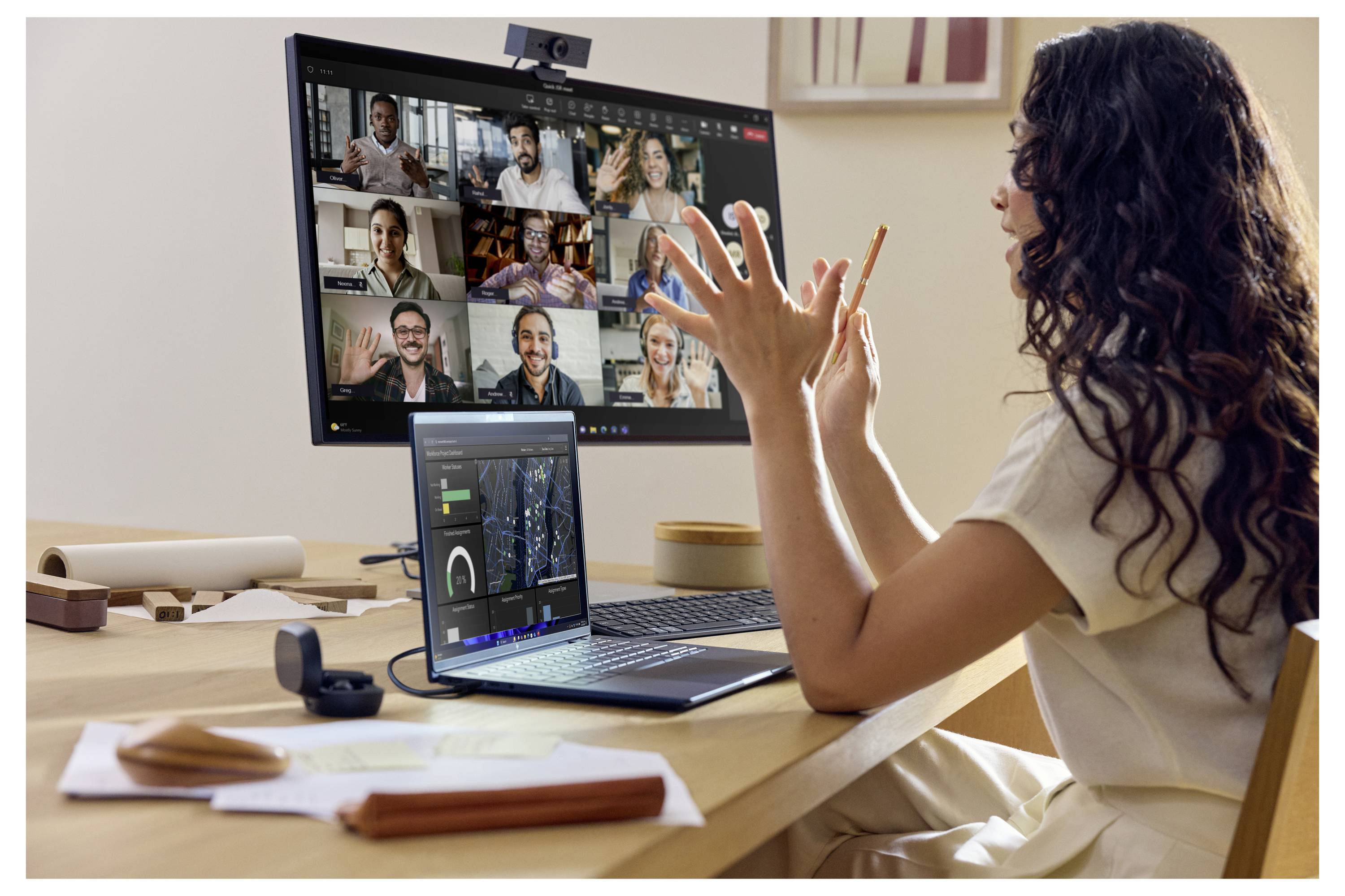 Frau bei einer Videokonferenz an einem Schreibtisch zu Hause, die auf einen Laptop und einen großen Monitor zeigt, auf denen ein Multi-Person-Meeting-Grid angezeigt wird.