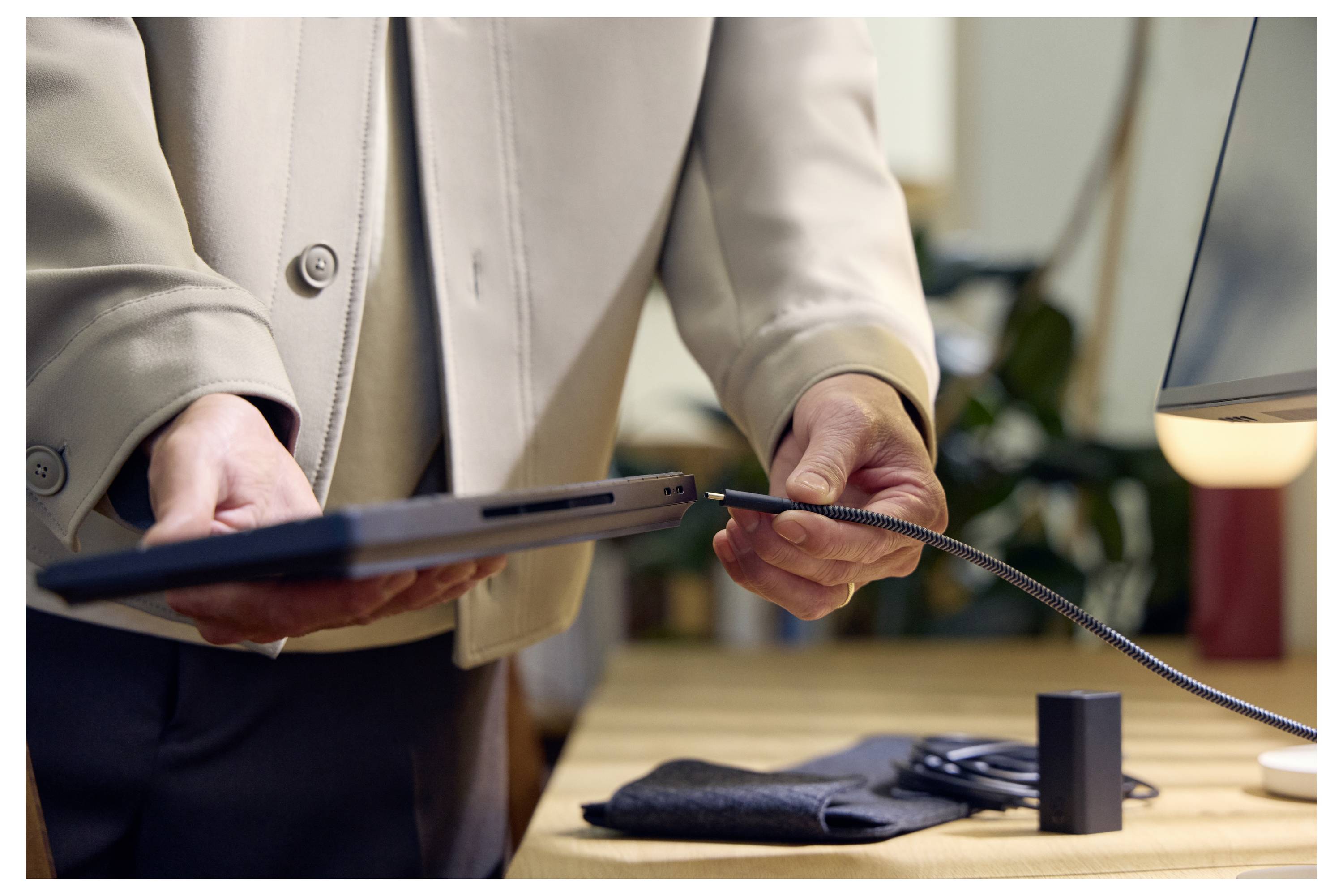 Eine Person in einem beigefarbenen Blazer verbindet ein Kabel mit einem Laptop an einem Schreibtisch, mit einer Lampe, einem Telefon und einem kleinen schwarzen Gerät in der Nähe.