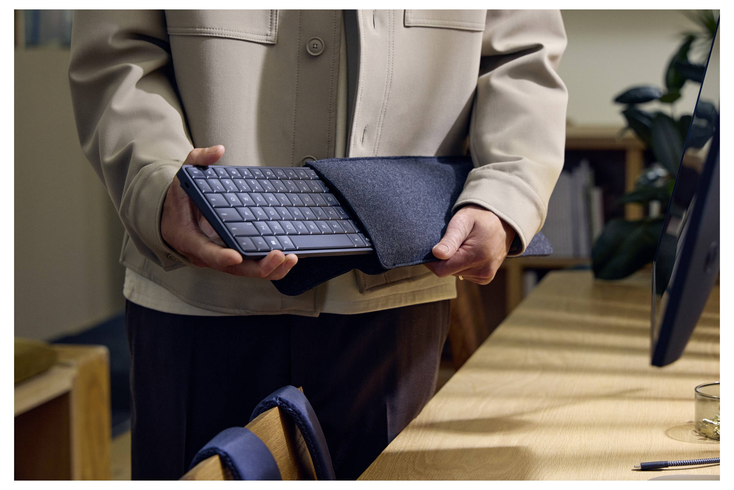 Eine Person hält ein Tablet in einer Tastaturhülle und steht neben einem Schreibtisch in einer Büroumgebung.