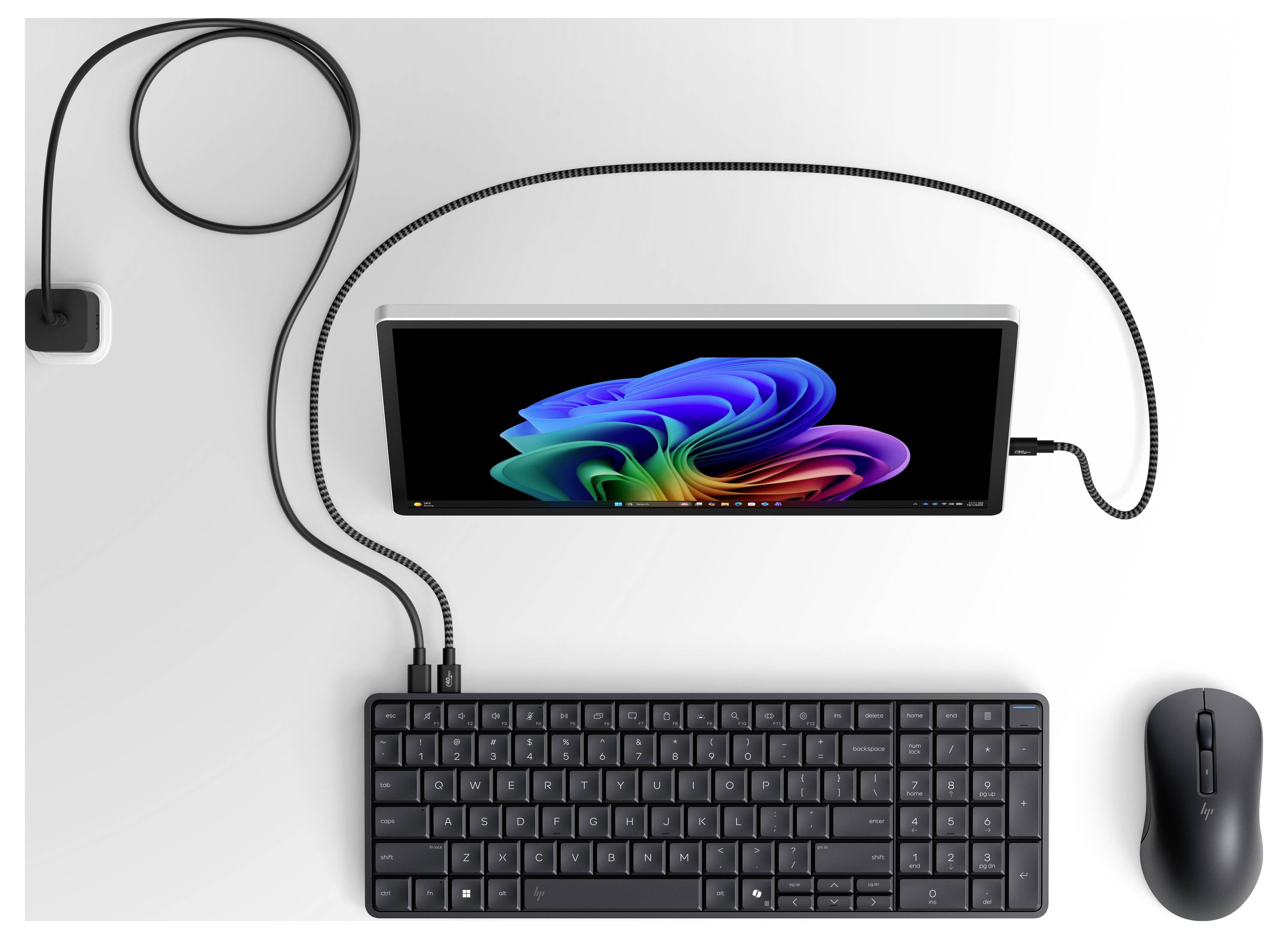 Draufsicht eines verkabelten Desktop-Setup mit einem zentralen Monitor, Tastatur, Maus und Netzteil, die durch Kabel auf einer weißen Oberfläche verbunden sind.
