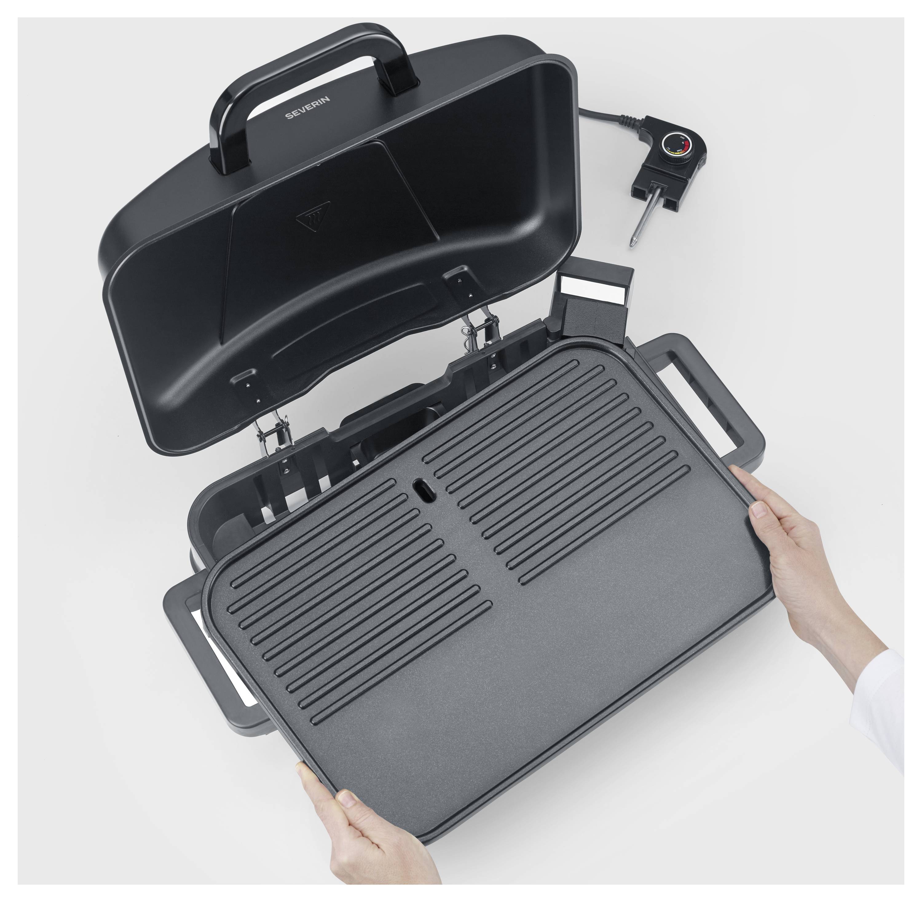 Hände öffnen einen schwarzen elektrischen Grill oder Grillplatte mit Scharnieren und zeigen die Kochplatte und den Deckel auf weißem Hintergrund.