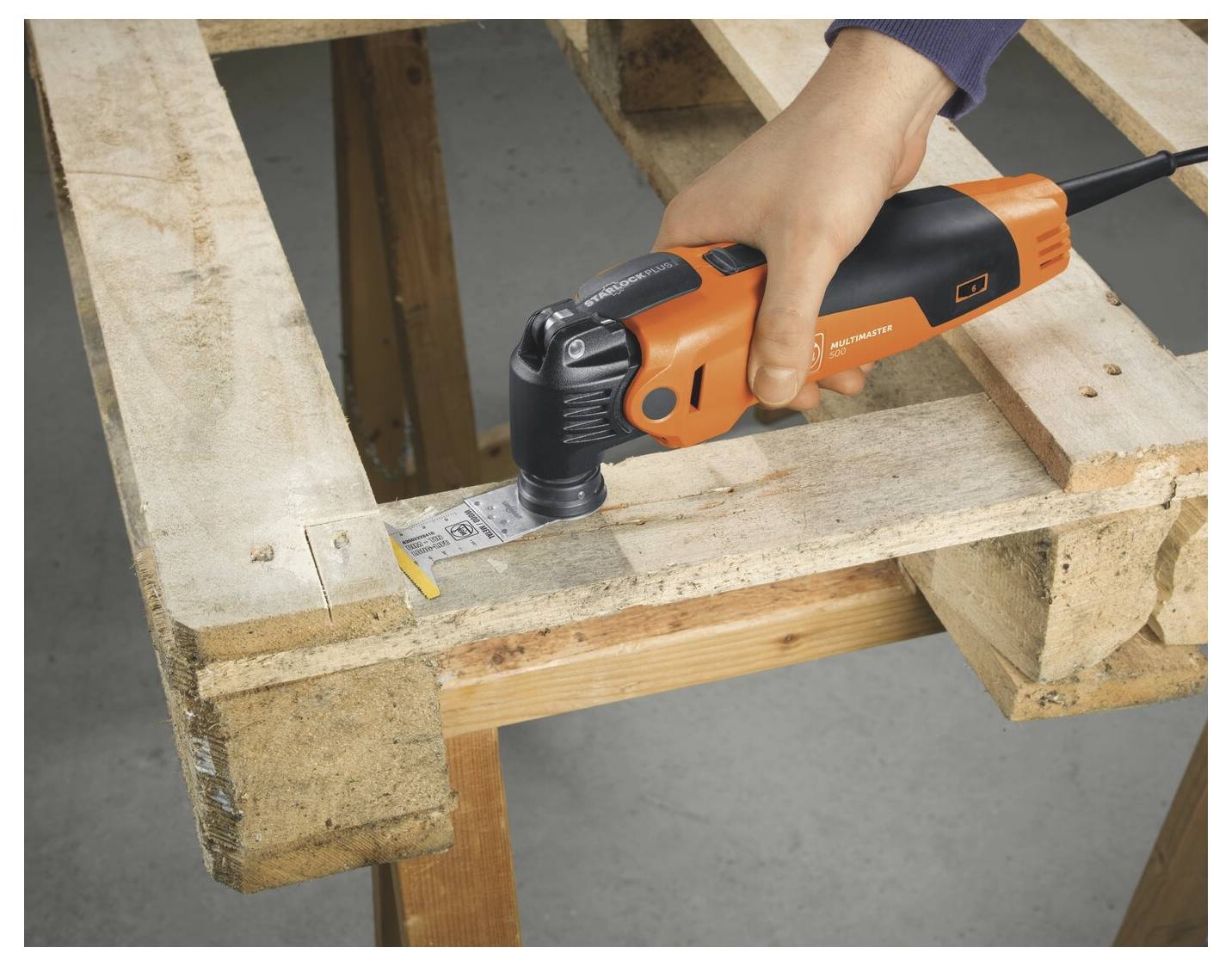 Hand, die ein oranges oszillierendes Multitool zum Schneiden einer Holzpalettendiele verwendet.