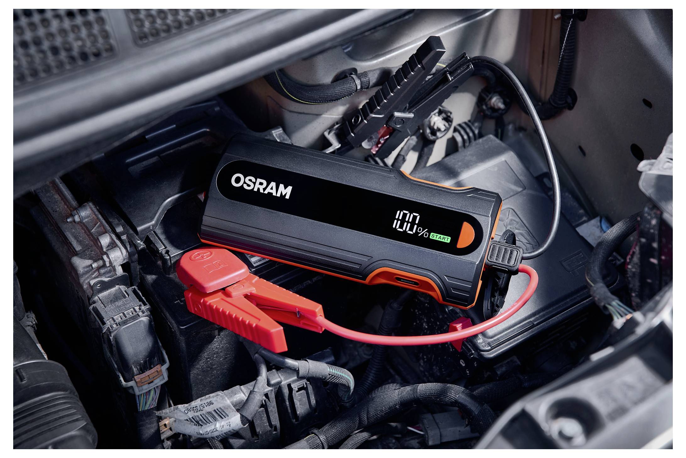 OSRAM portable car jump starter connected to a vehicle battery with red and black clamps in the engine bay, showing 100% charge.