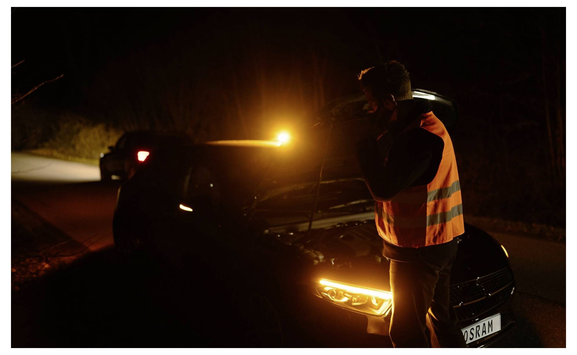 Eine Person in einer Warnweste inspiziert nachts ein liegengebliebenes Auto, das von den Scheinwerfern des Autos und einem bernsteinfarbenen Warnlicht beleuchtet wird.