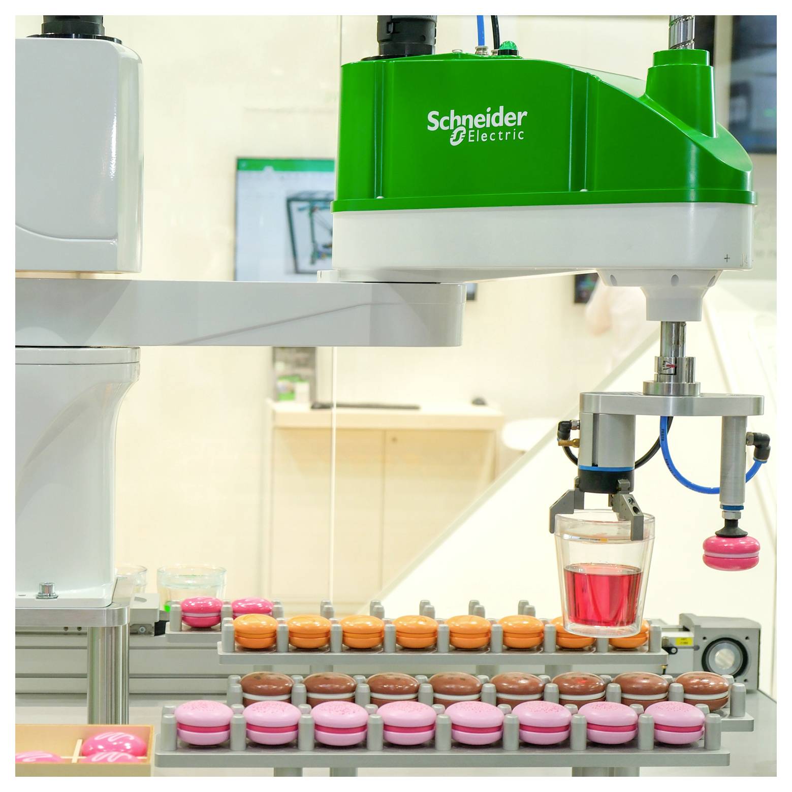Ein Industrieroboterarm von Schneider Electric bewegt eine rot gefüllte Tasse über Förderbänder mit bunten runden Behältern in einer Fabrik oder einem Labor.