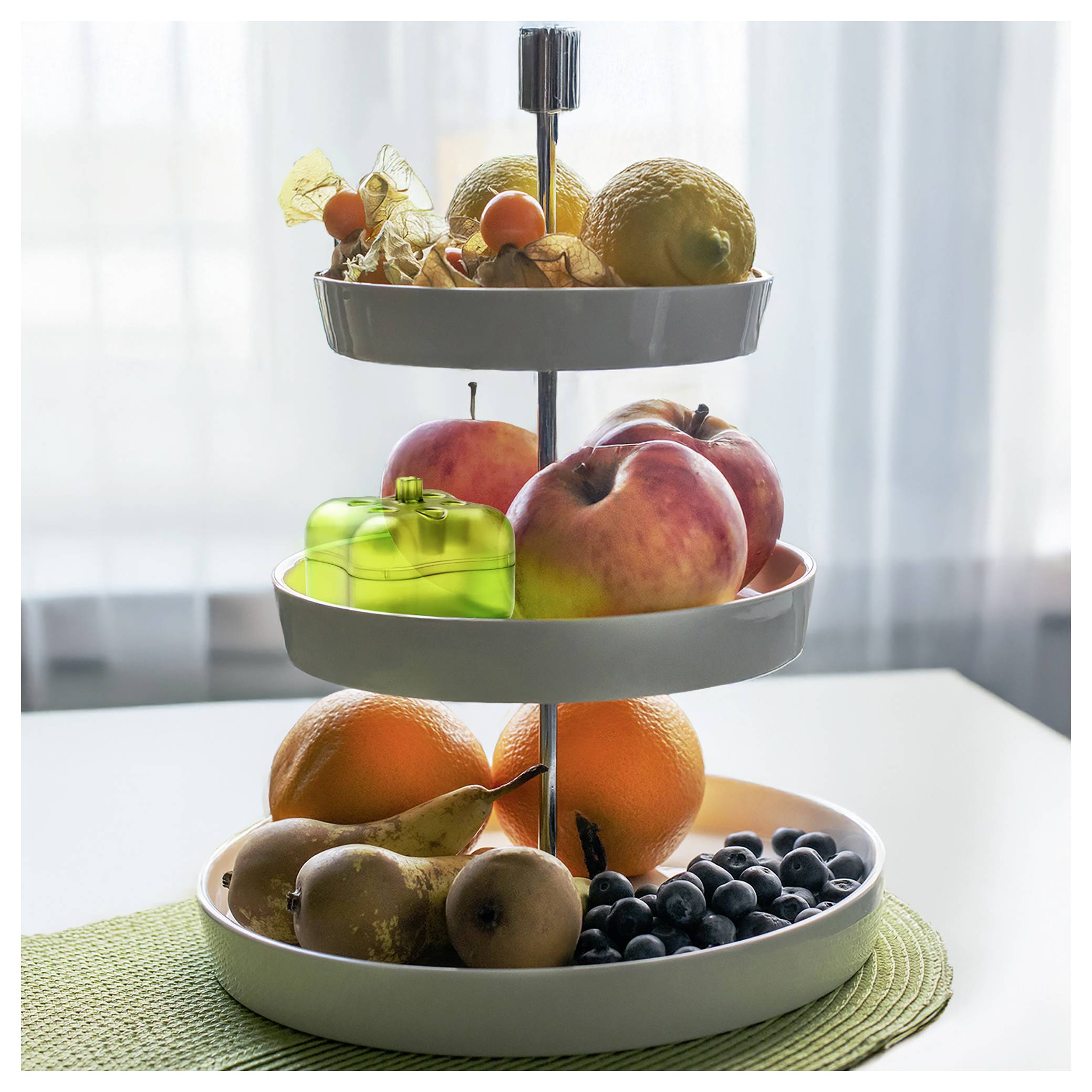 Dreistöckiger Obststand mit gemischtem frischem Obst, einschließlich Orangen, Äpfeln, Pfirsichen, Kiwis, Blaubeeren und Trockenobst auf einem Tisch neben einem Fenster.