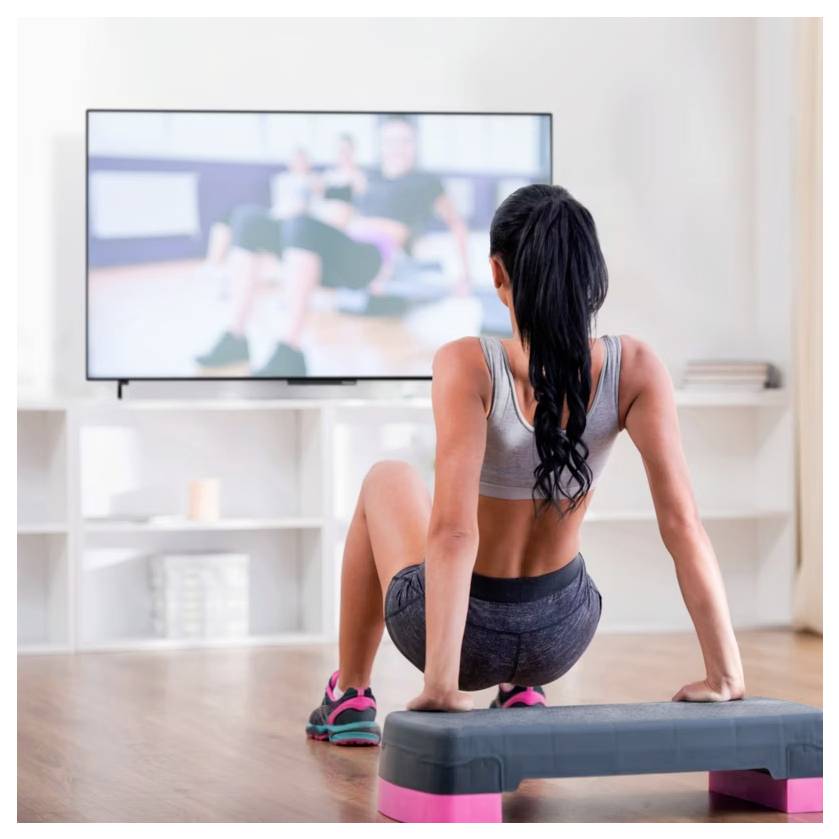 Frau trainiert zu Hause, während sie sich ein Trainings-Video auf dem Fernseher ansieht, sitzt auf einer Aerobic-Stufe in einem hellen Wohnzimmer.