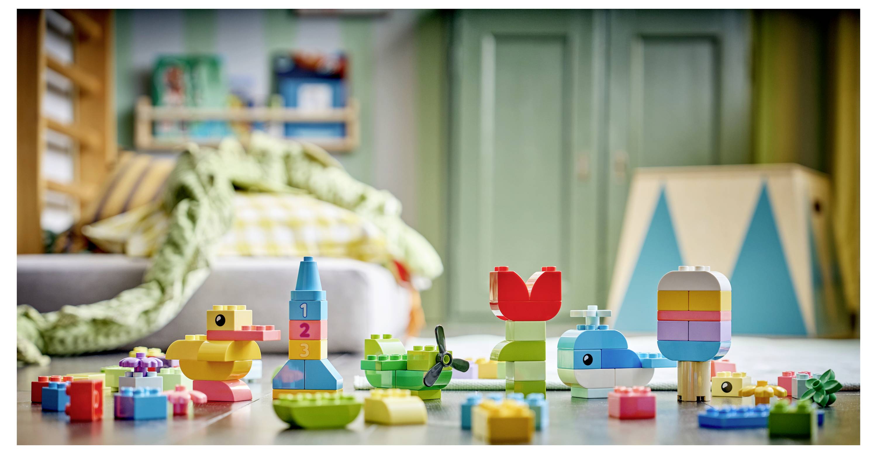 Bunte Spielbausteine auf dem Boden vor einem unscharfen Schlafzimmer angeordnet, mit einem Bett und einem Spielzelt im Hintergrund.