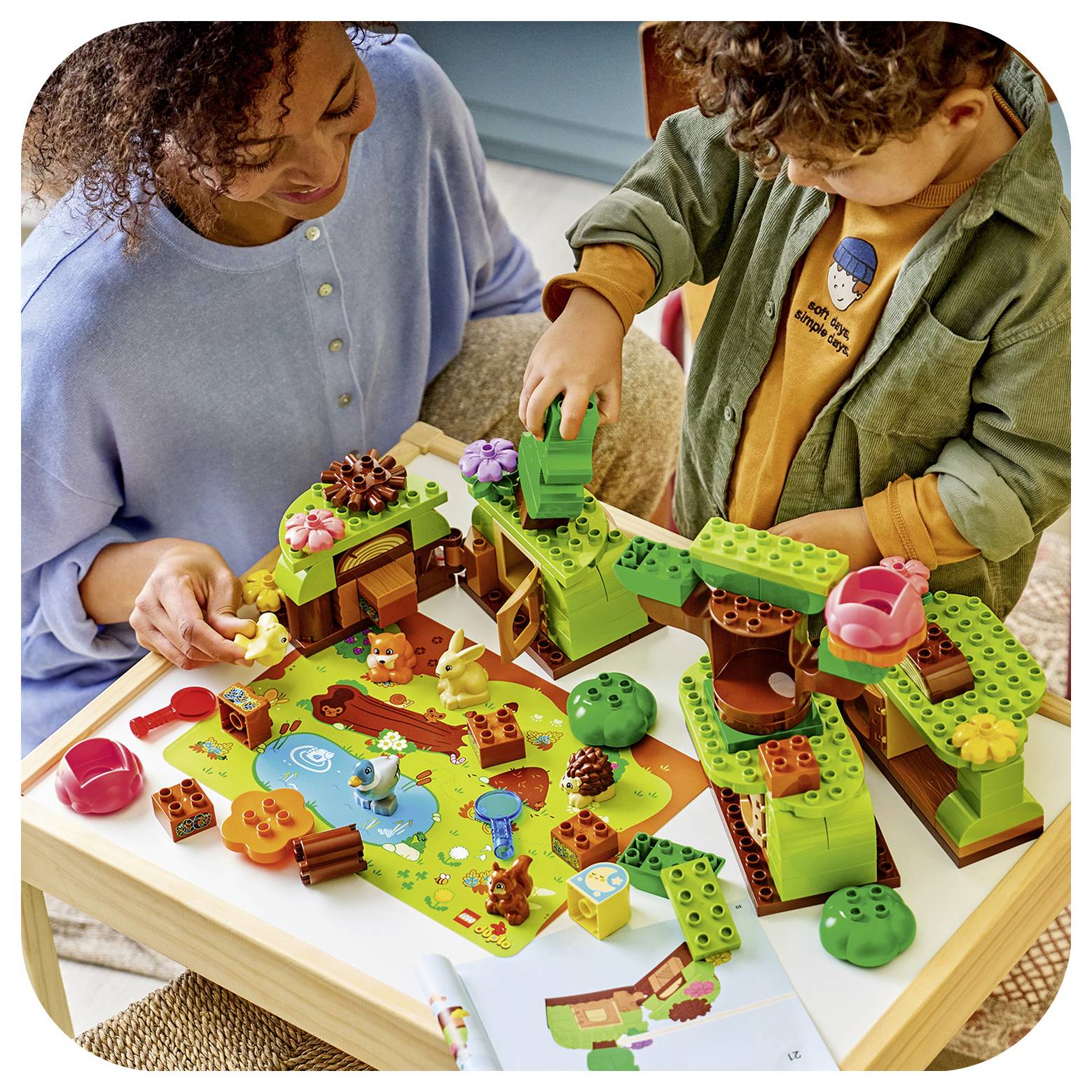 Ein Erwachsener und ein Kind spielen mit einem farbenfrohen LEGO-ähnlichen Waldbauset auf einem Tisch, bauen Bäume, Brücken und Tierfiguren auf einer illustrierten Spielmatte zusammen.