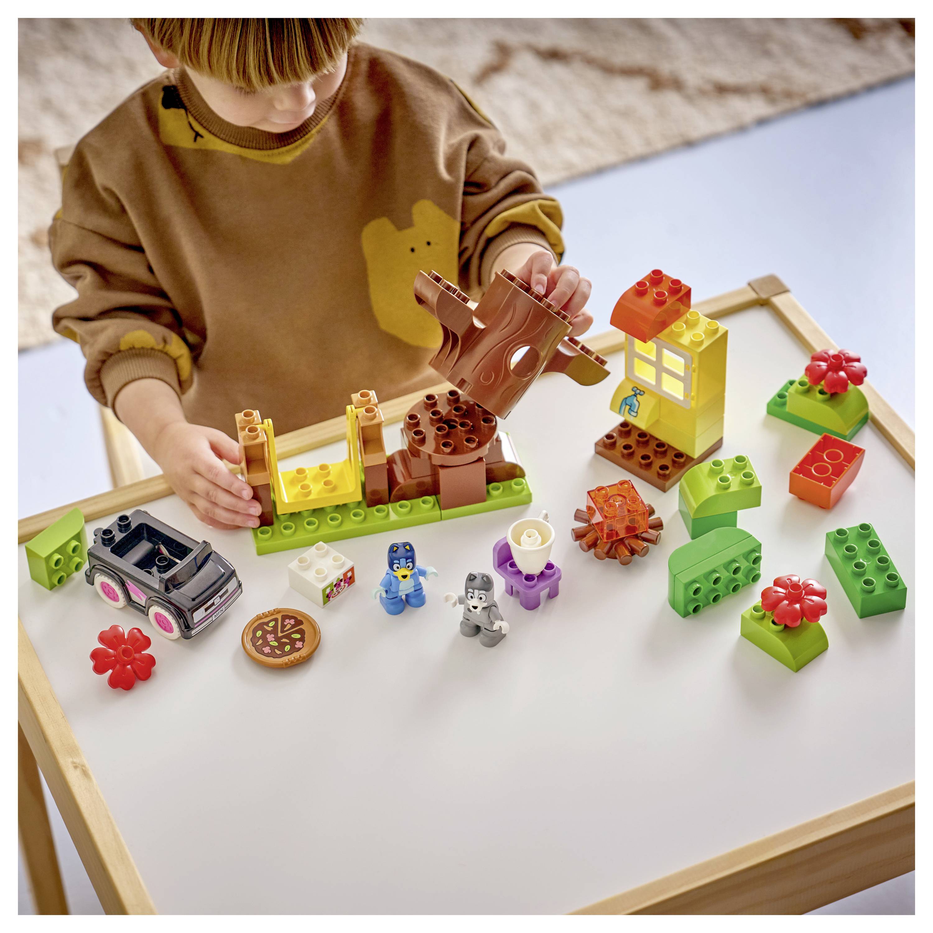 Kind spielt mit bunten Bauklötzen und kleinen Spielfiguren auf einem Tisch und baut eine Spielszene mit einem Haus und einem Baum zusammen.