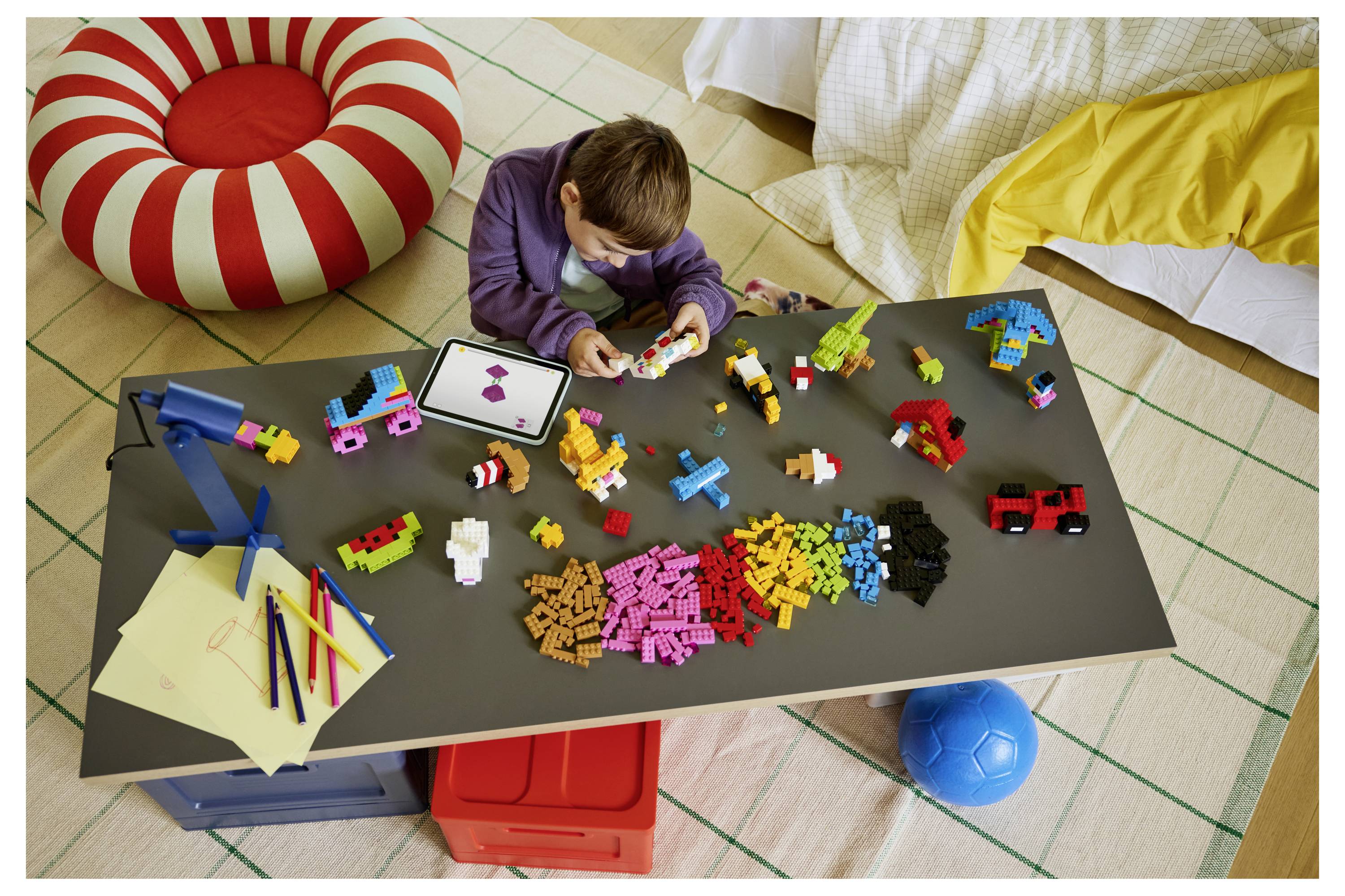 Kind, das mit farbigen Bausteinen an einem niedrigen Tisch spielt, umgeben von Spielzeug und Kissen in einem Spielzimmer.