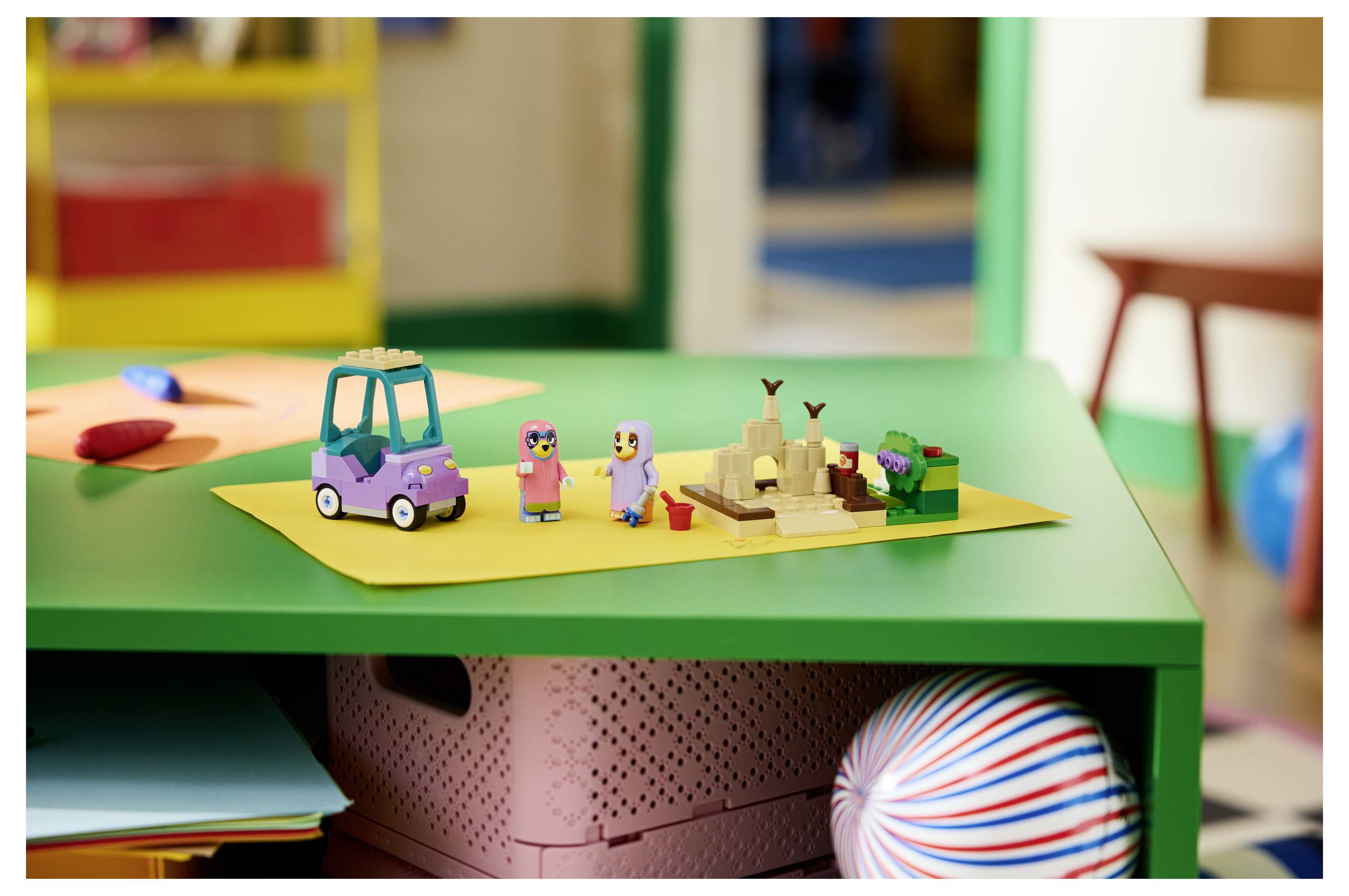 Spielzeugszene auf einem grünen Tisch mit einem lila Auto, zwei kleinen Figuren und einer Sandburg auf gelbem Papier, was ein verspieltes Kindersetup suggeriert.