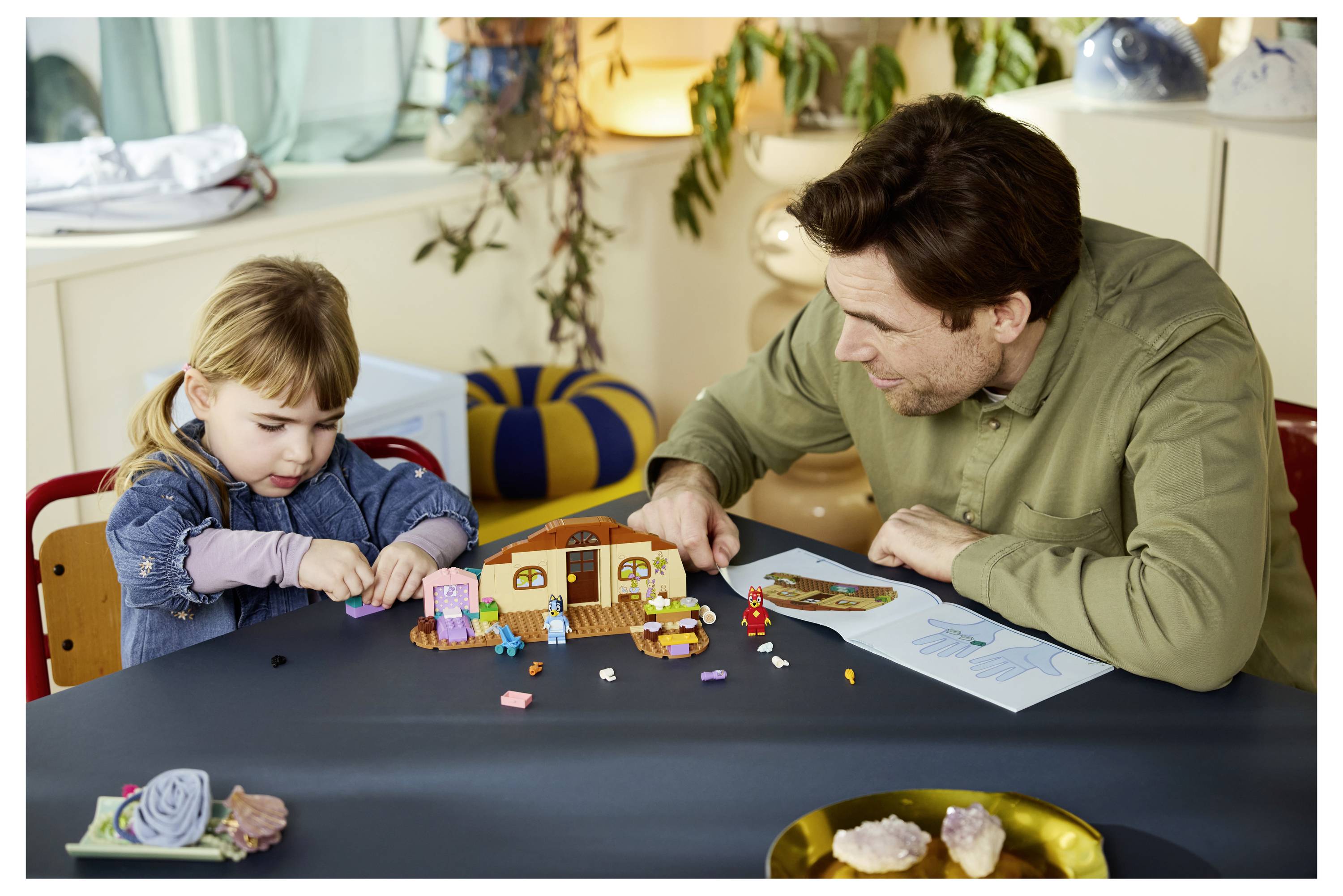 Ein Erwachsener und ein kleines Kind spielen zusammen mit einem LEGO-ähnlichen Hausset an einem Tisch und folgen dabei gemeinsam einer Anleitung.