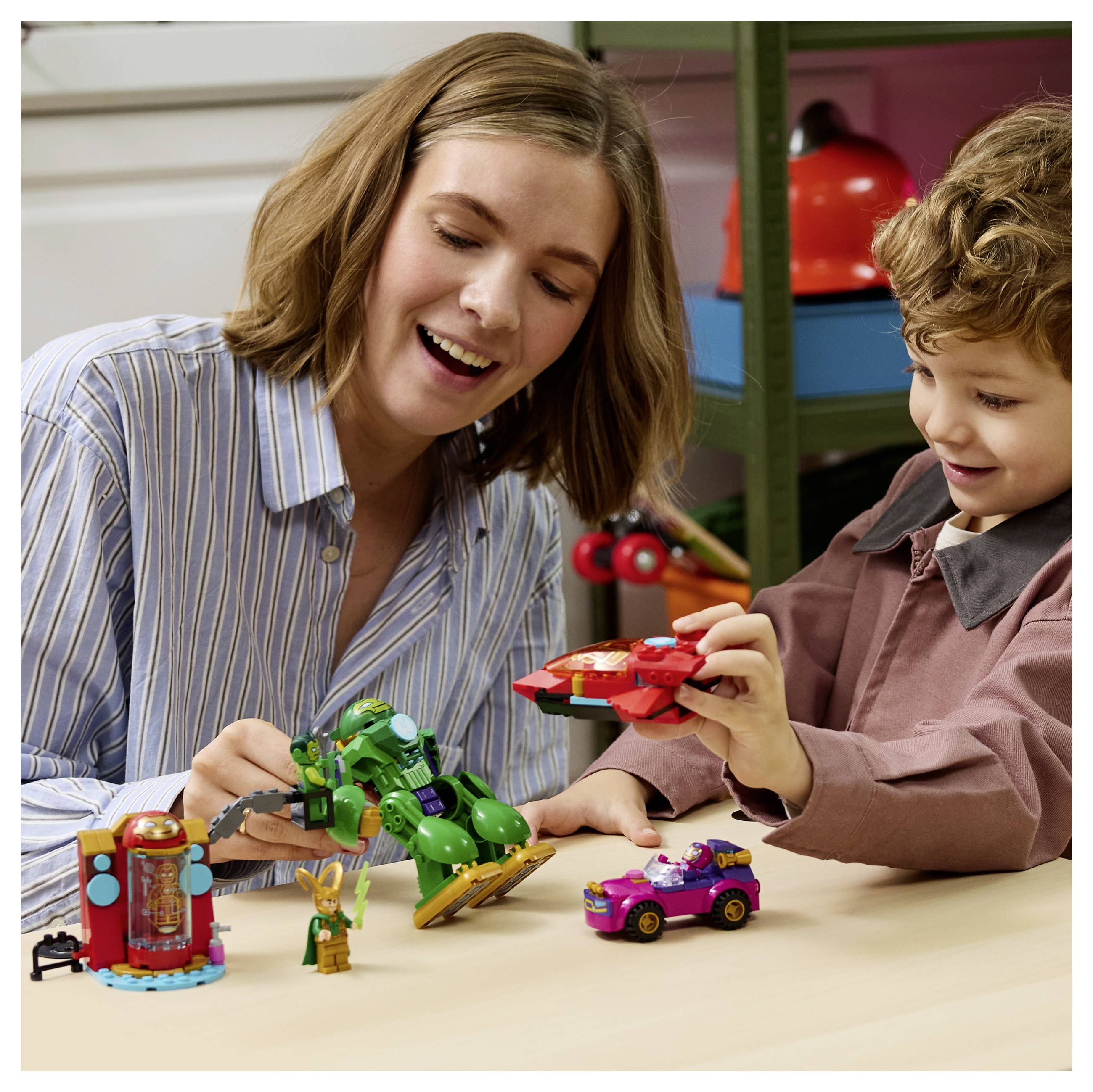 Frau und kleiner Junge lächeln, während sie mit bunten Spielzeugfahrzeugen und Figuren an einem Tisch im Spielzimmer spielen.