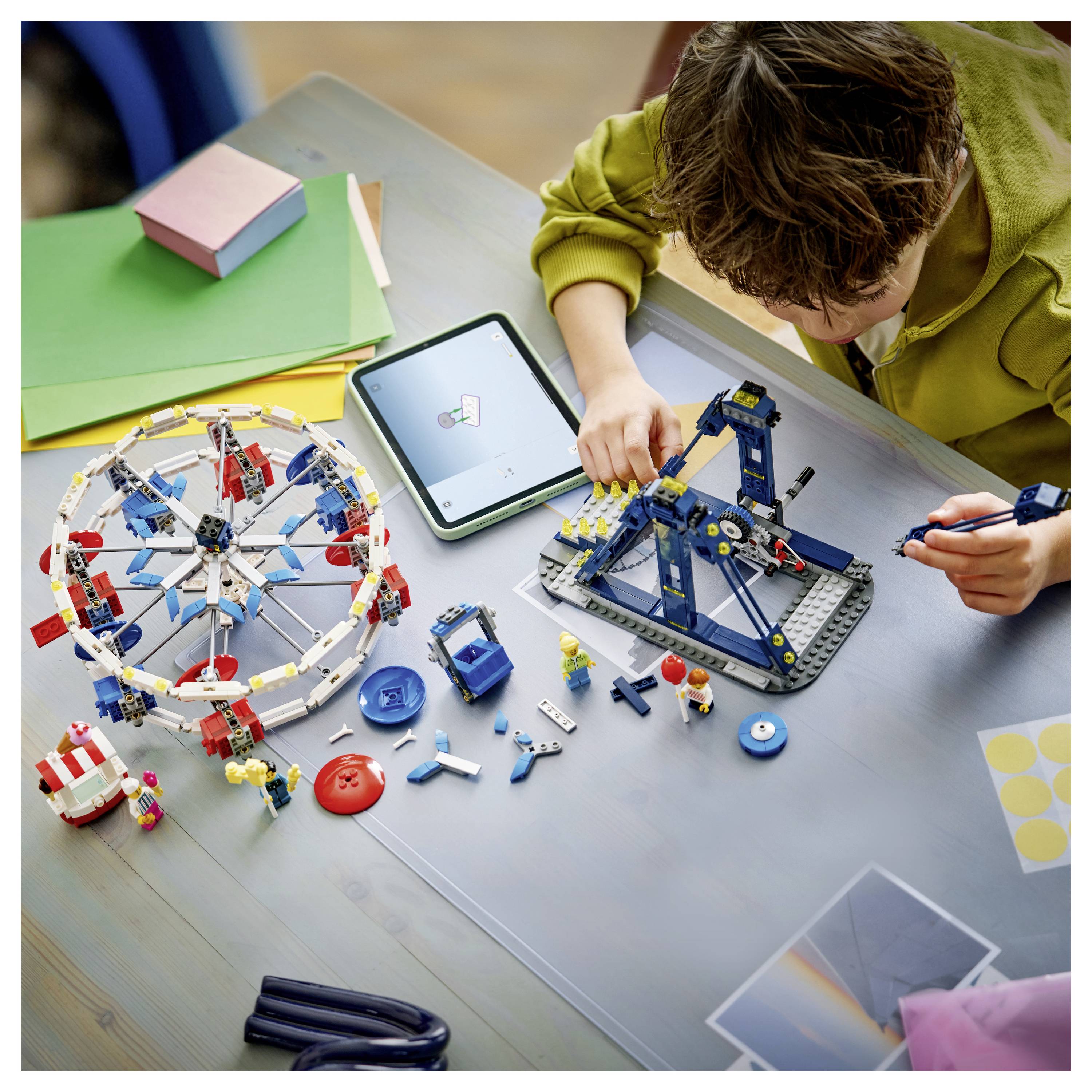 Ein Kind baut ein LEGO-Riesenrad und einen Kran an einem Tisch, mit verstreuten Teilen, einem Tablet und anderen Bastelmaterialien herum.
