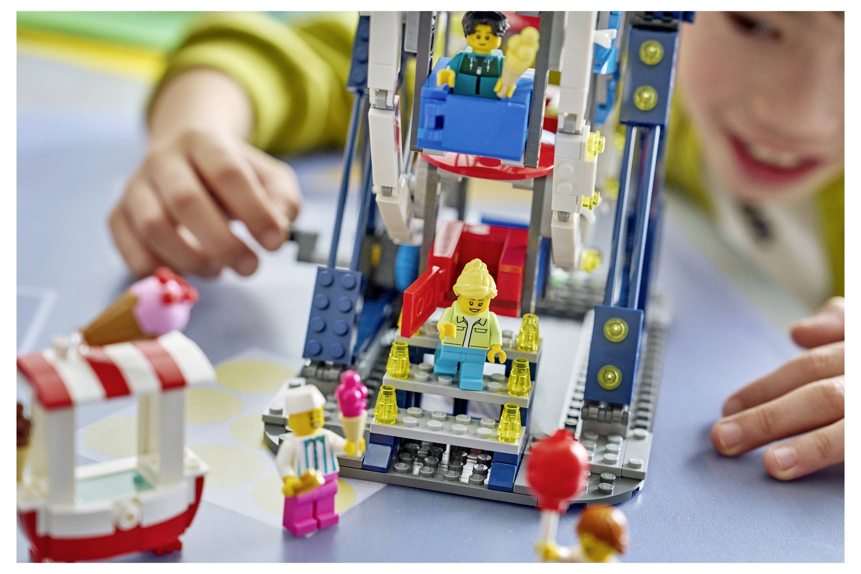 Kind spielt mit einem farbenfrohen LEGO-Fahrgeschäft oder Riesenrad, mit mehreren sichtbaren Minifiguren in einer verspielten Innenszene.