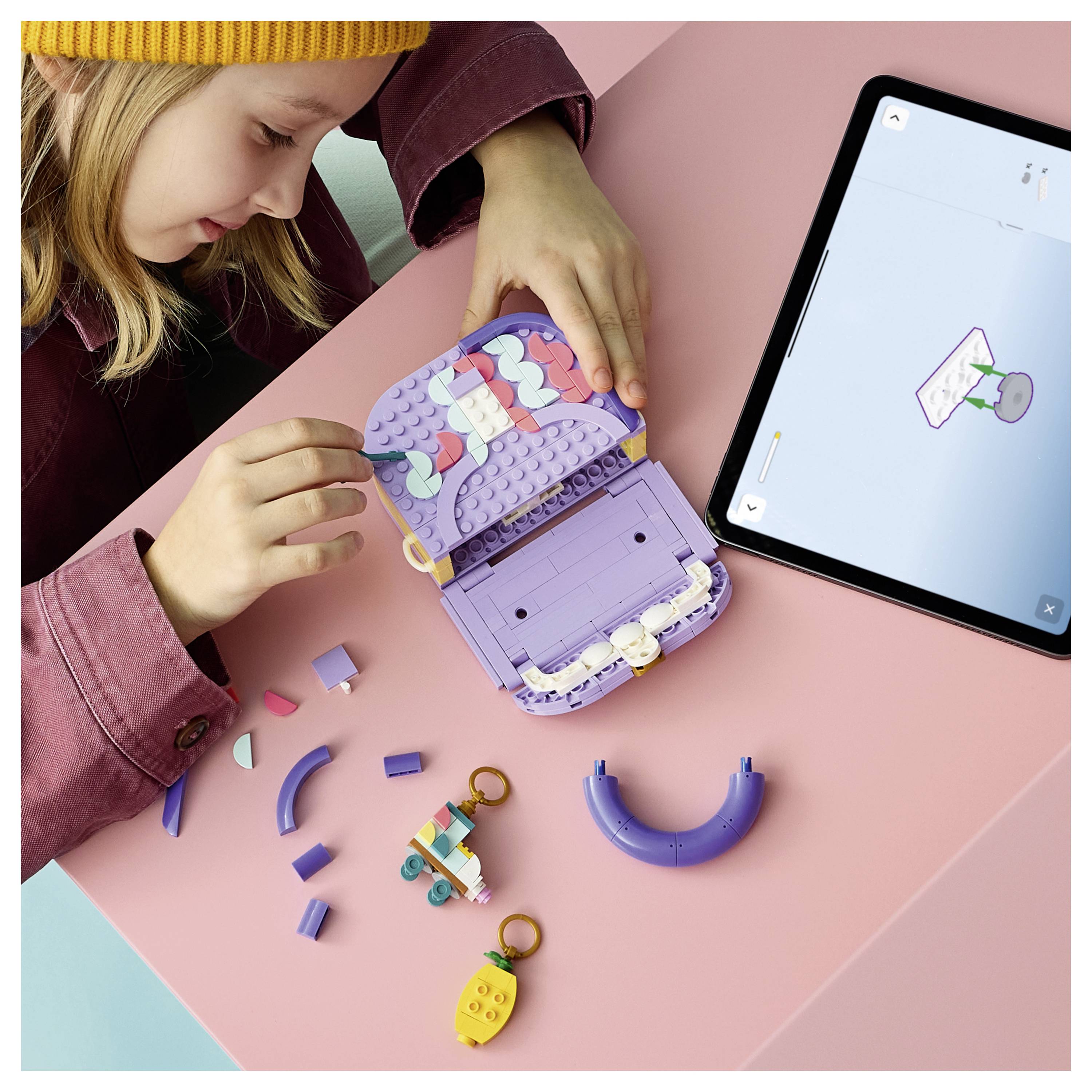 Kind baut eine violette LEGO-ähnliche Spielzeugtasche auf einem rosa Tisch zusammen, während es Anweisungen auf einem Tablet neben verstreuten Teilen befolgt.