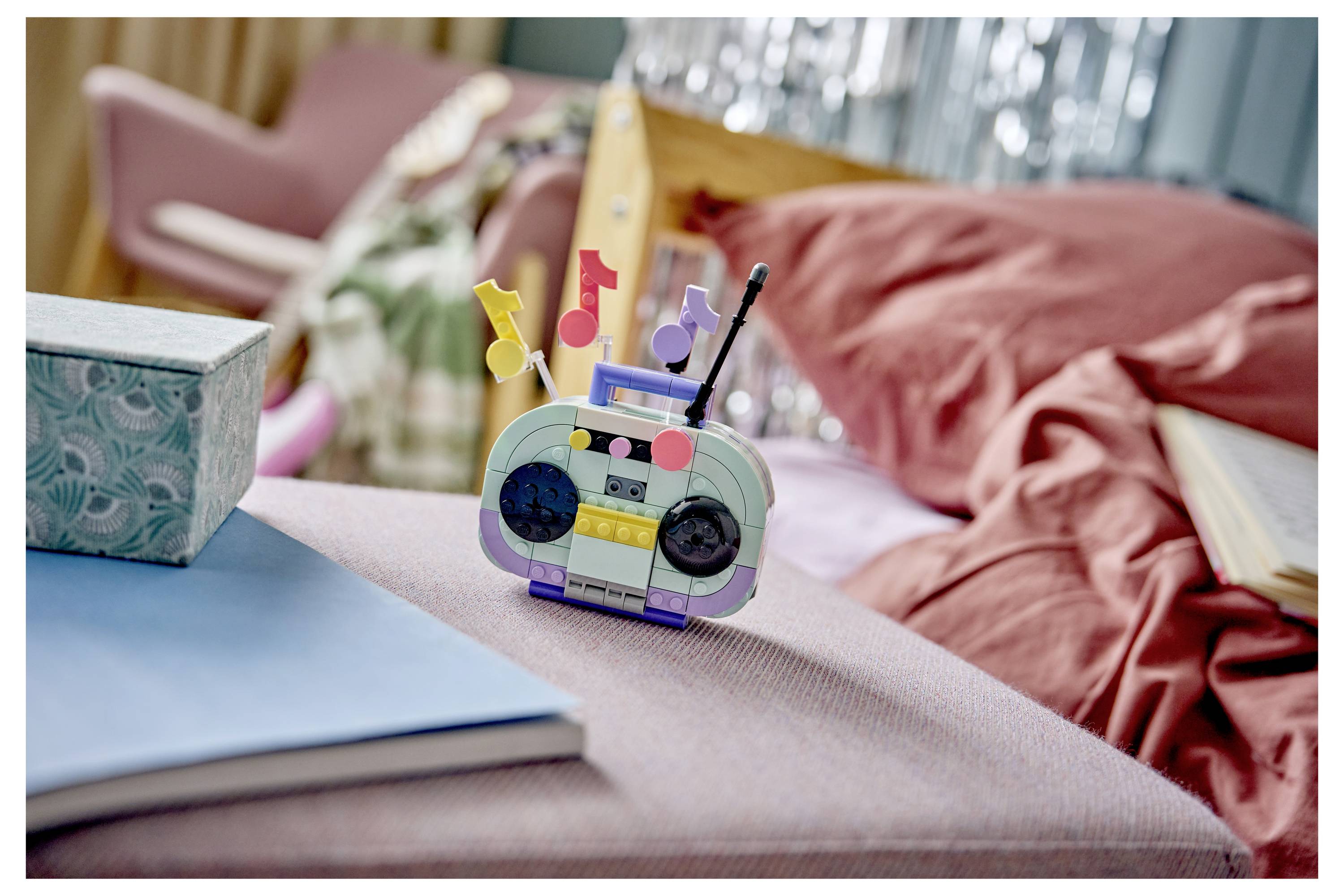 LEGO-ähnlicher Musikroboter-Spielzeug auf einem rosa Sofa neben einem blauen Buch und einer Taschentuchbox, mit einem gemütlichen Wohnzimmer-Hintergrund.