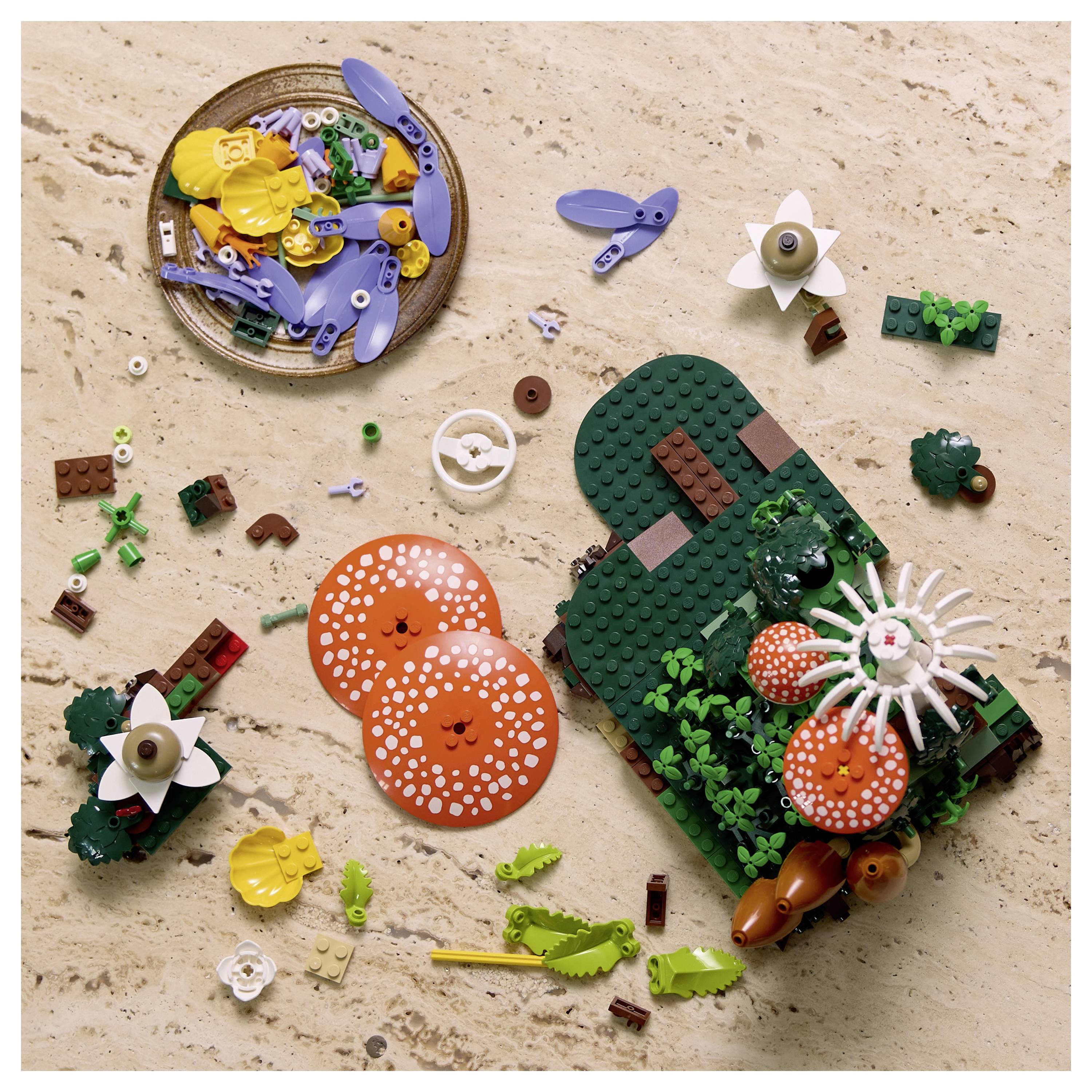 Draufsicht auf ein LEGO-ähnliches Diorama auf einer sandigen Oberfläche, das eine grüne Insel mit Pflanzen, orangefarbenen Pilzen, kleine Figuren und einen Korb mit verschiedenen Teilen zeigt.