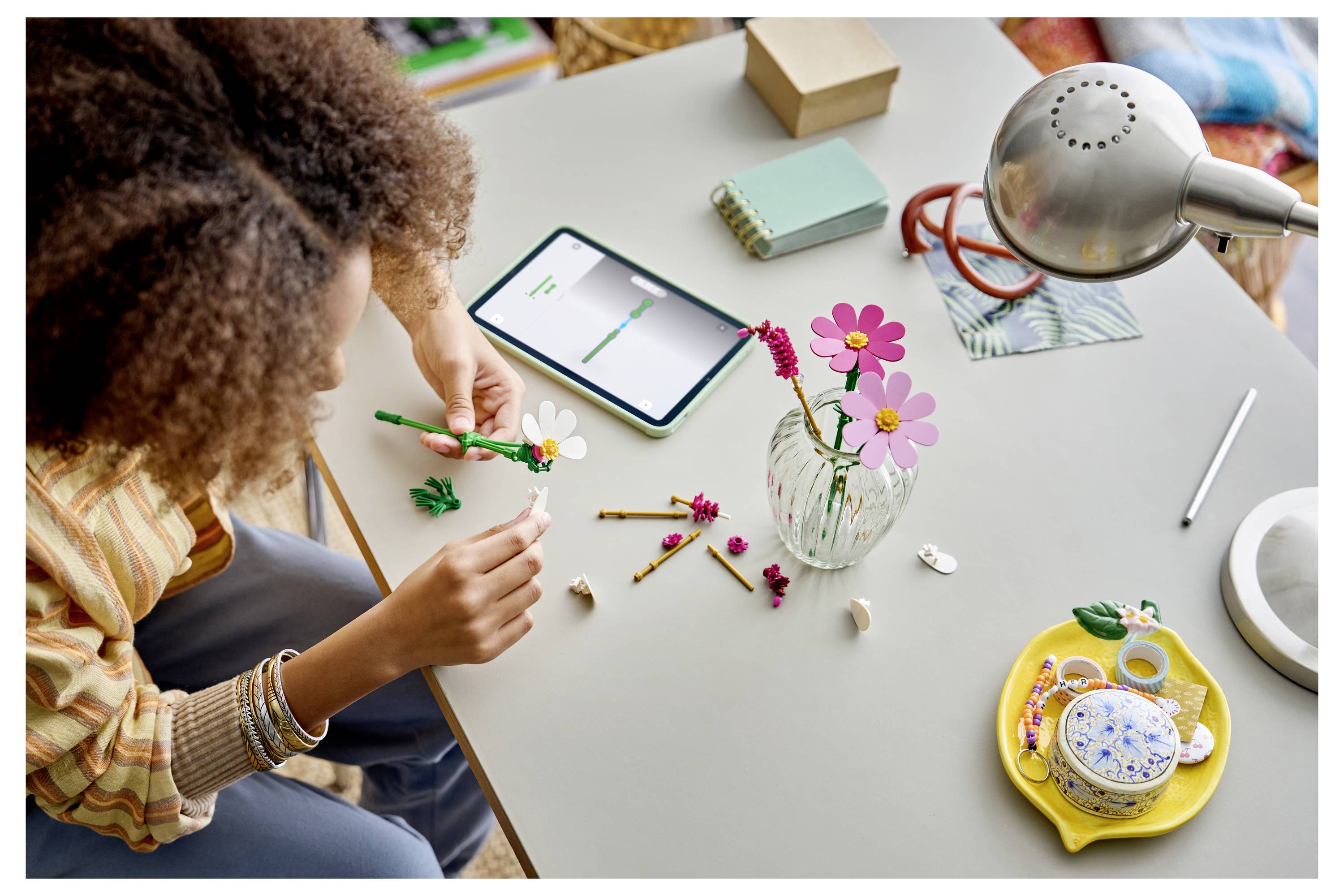 Eine Person, die einen Blumenstrauß an einem Tisch arrangiert, nutzt dabei ein Tablet als Referenz, mit Werkzeugen, Blumen und einer Vase in der Nähe.