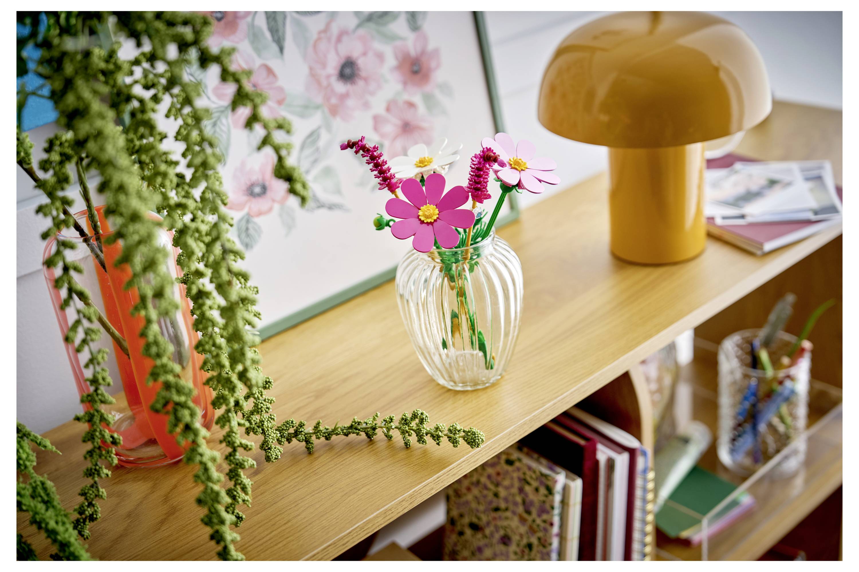Dekoratives Regal mit einer gestreiften Vase mit rosa und magentafarbenen Blumen, hängender grüner Pflanze, orangefarbener Lampe und Büchern in einem hellen Raum.