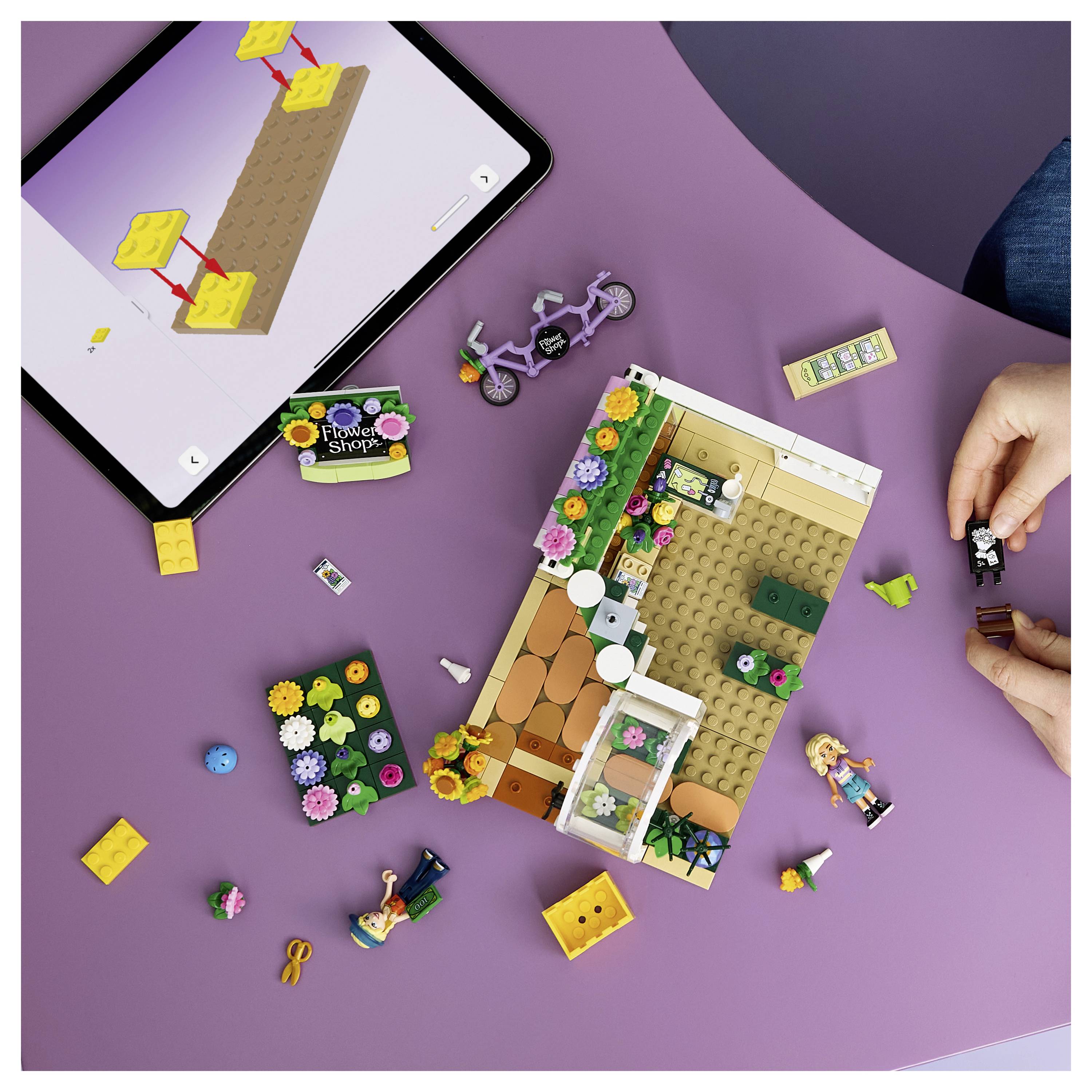 Hände, die ein LEGO-ähnliches Blumenladen-Set neben einem Tablet mit Bauanweisungen zusammenbauen, mit losen Teilen und einem Fahrrad auf einer violetten Oberfläche.