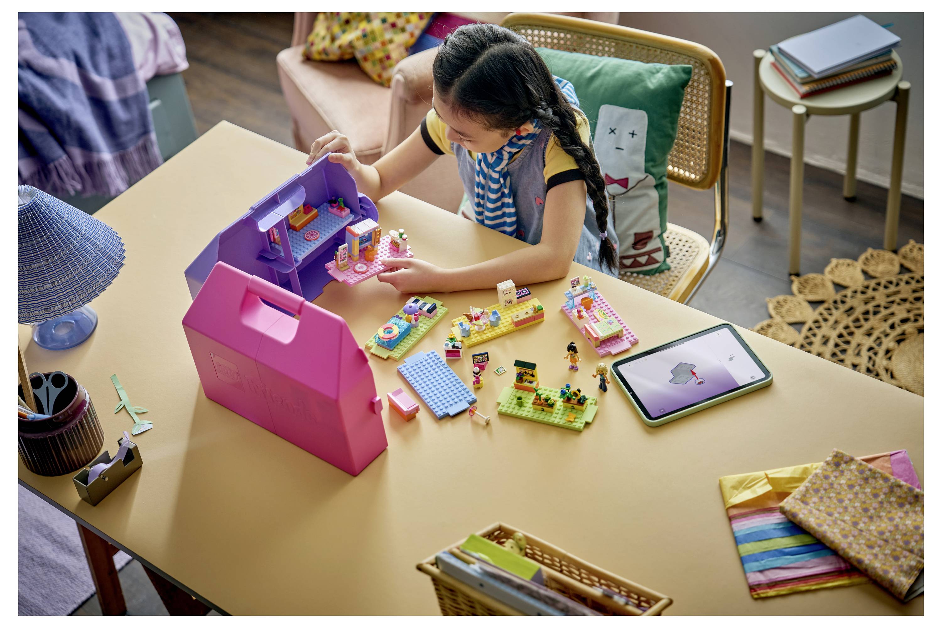 Ein Mädchen spielt mit einem Spielzeug-Puppenhaus und Bausteinen an einem Tisch, mit einem Tablet daneben und bunten Bastelmaterialien in der Nähe.