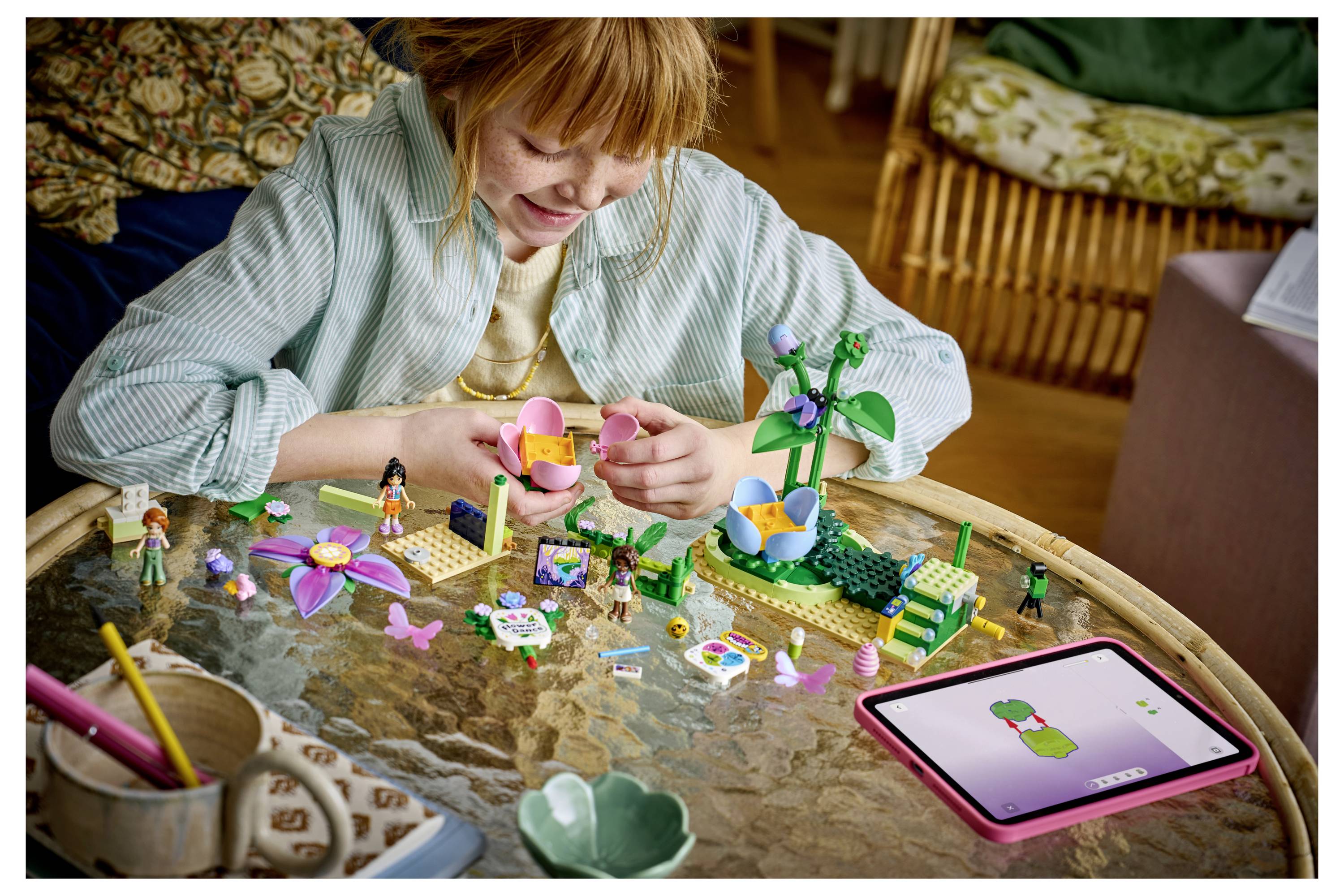 Mädchen baut einen bunten LEGO-Blumengarten auf einem Tisch auf, mit einem Tablet in der Nähe, das eine Blumenmodell-Anleitung zeigt.
