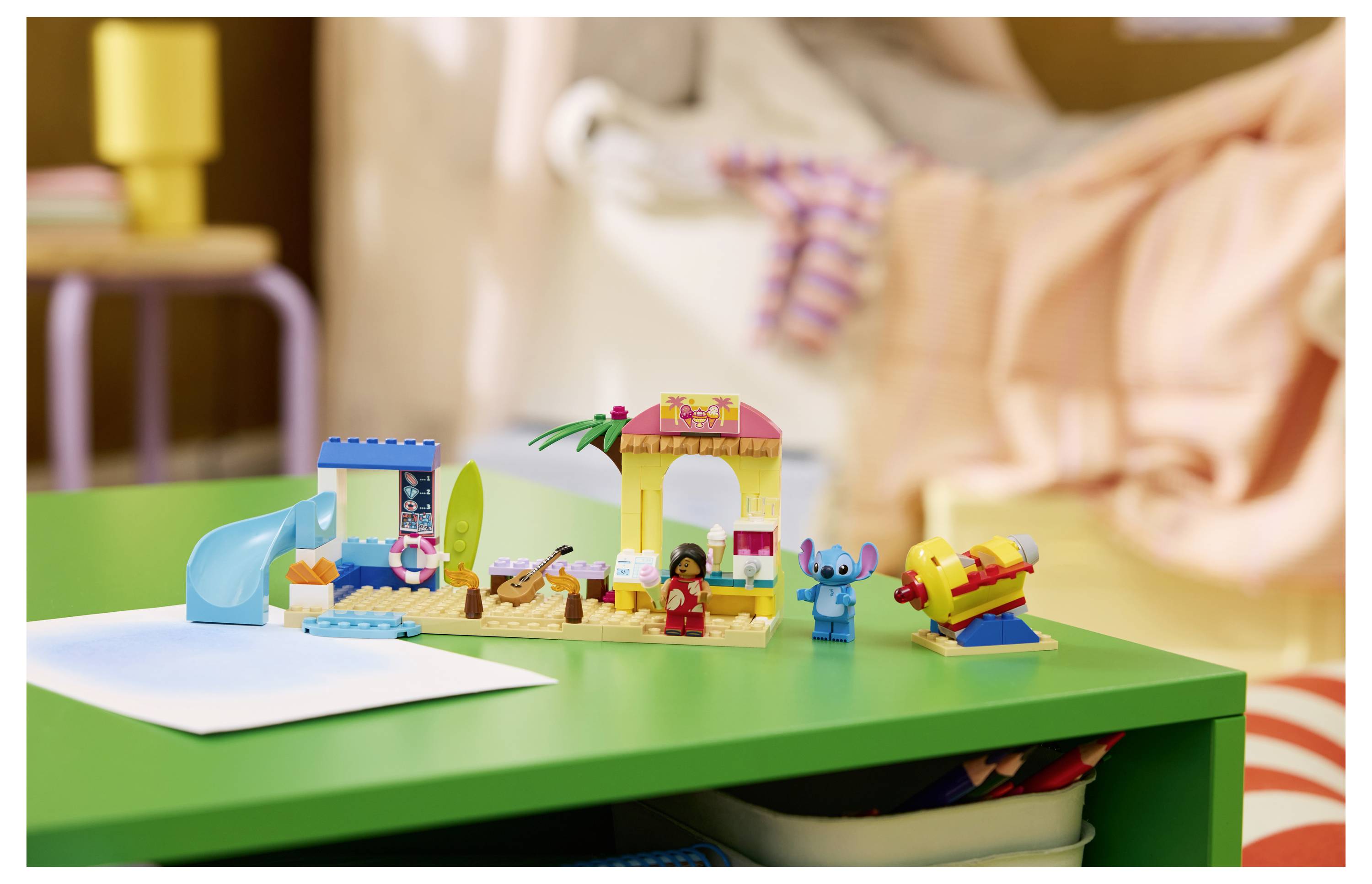 Bunte Spielbausteine auf einem grünen Tisch, darunter eine Rutsche, ein kleines Haus, Figuren und ein gelbes Flugzeug, mit einem unscharfen Kind im Hintergrund.