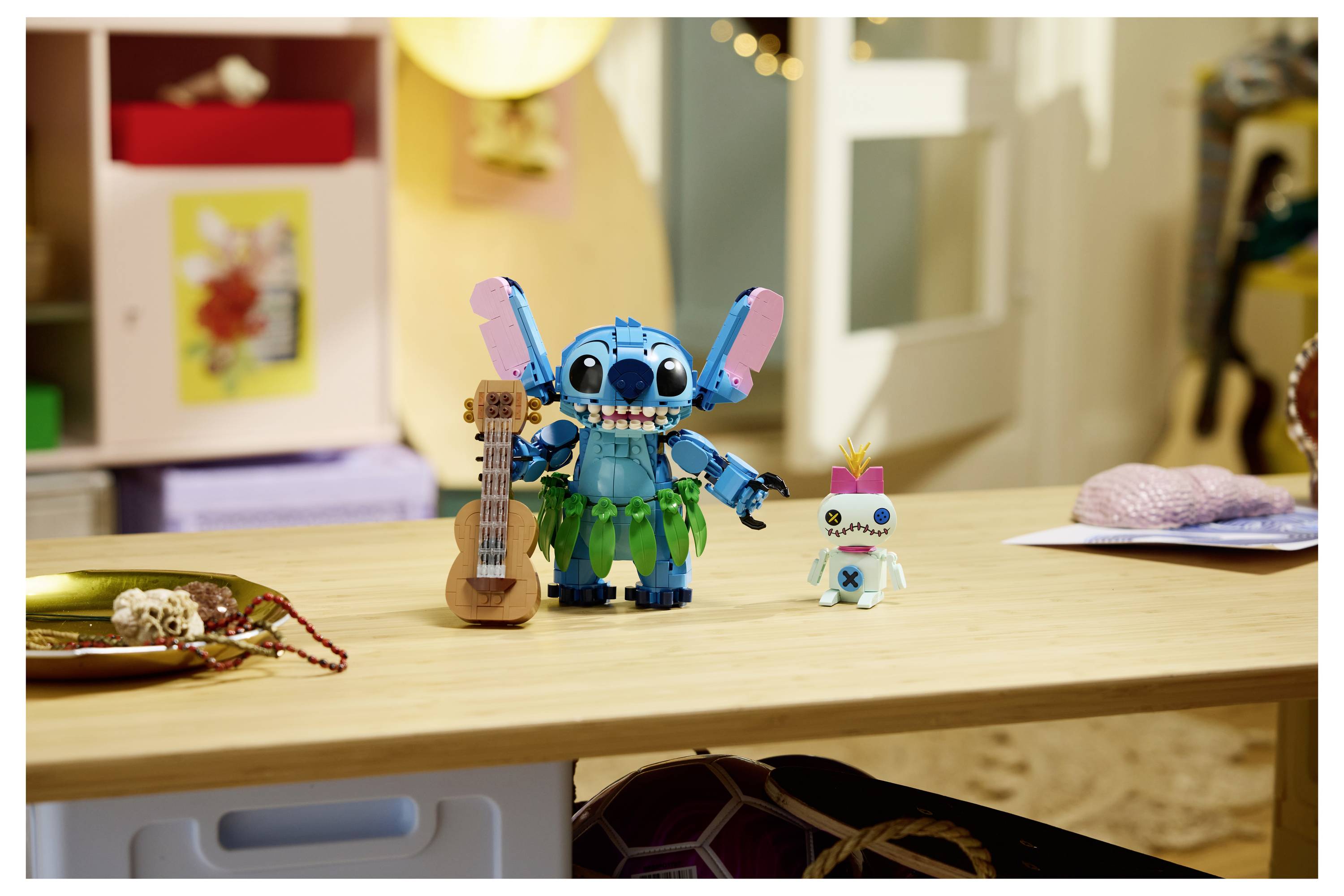 Zwei Spielfiguren auf einem Schreibtisch, darunter eine blaue Stitch-ähnliche Figur mit einer Ukulele und eine kleinere weiße Figur, in einer Raumkulisse.